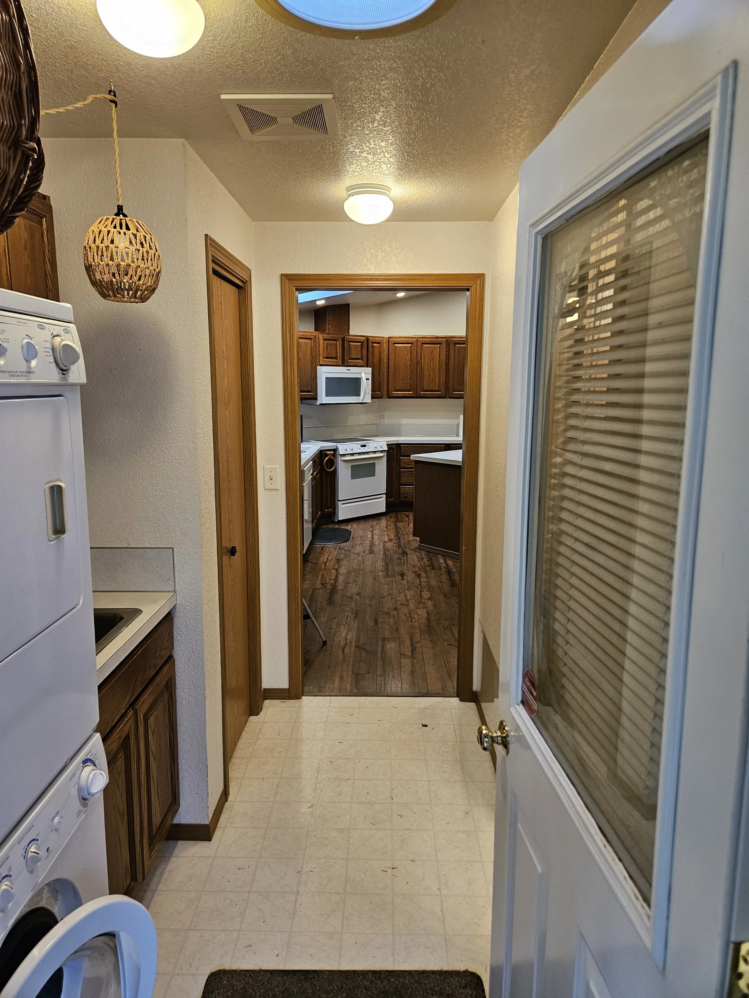 View from the entrance of a kitchen showing appliances, wooden cabinets, a microwave, and a stove with a walk-in pantry doorway leading to a room with dark wood flooring and cabinetry.