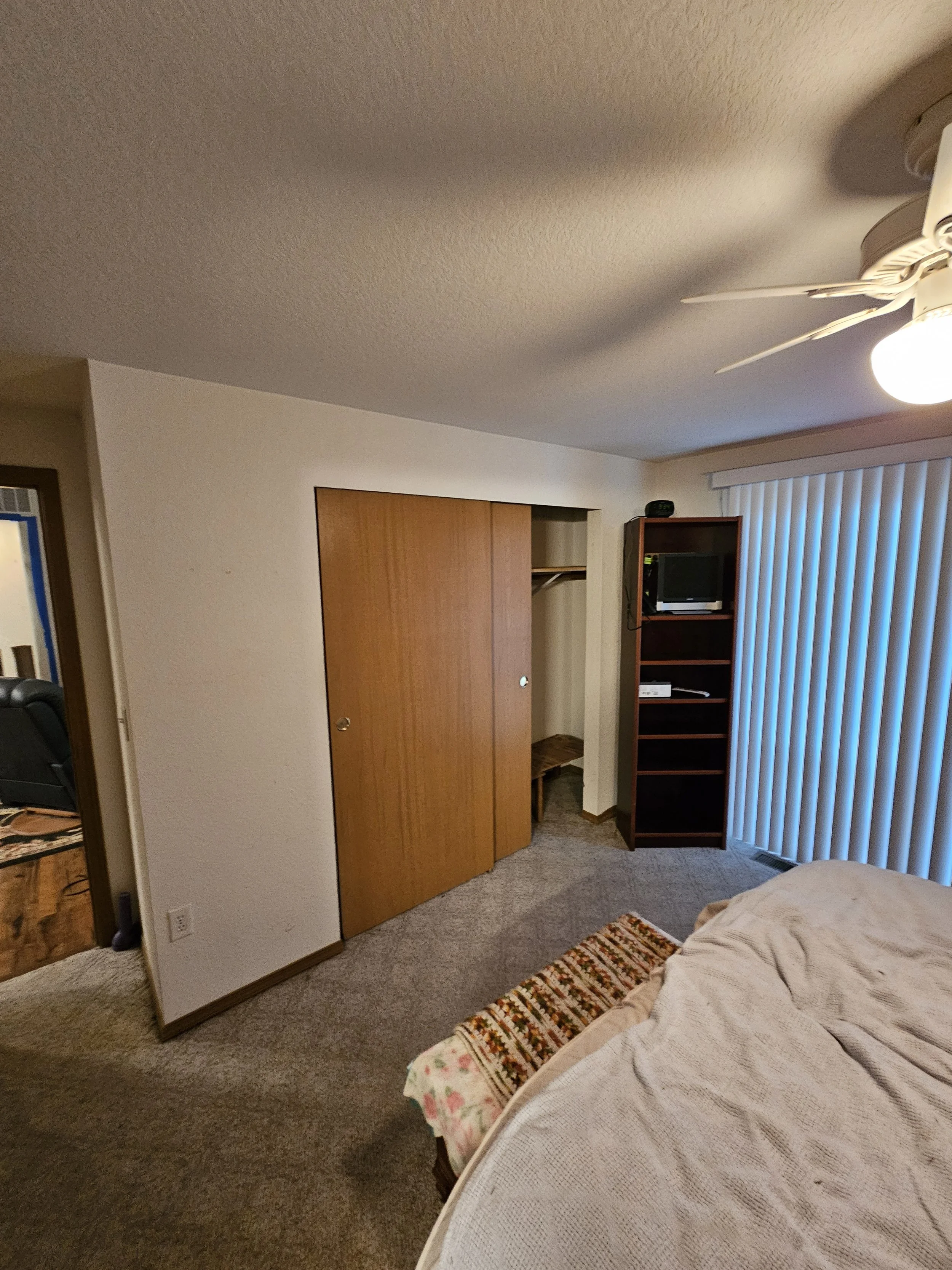 A bedroom with a bed, a carpeted floor, a sliding glass door with vertical blinds, a wooden closet door, a small TV on a wooden stand, a ceiling fan with lights, and part of a floral bedspread.