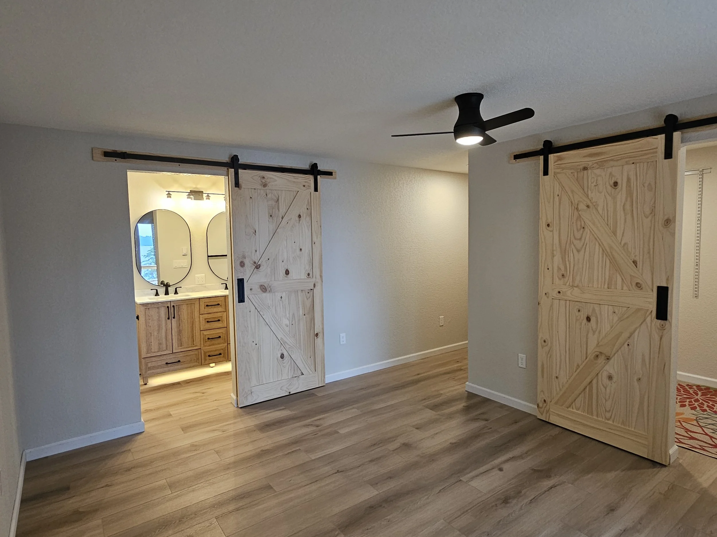 Interior of a room with two sliding barn doors on black tracks, an open doorway leading to a bathroom with two oval mirrors and a vanity, light-colored walls, wood flooring, and a black ceiling fan.