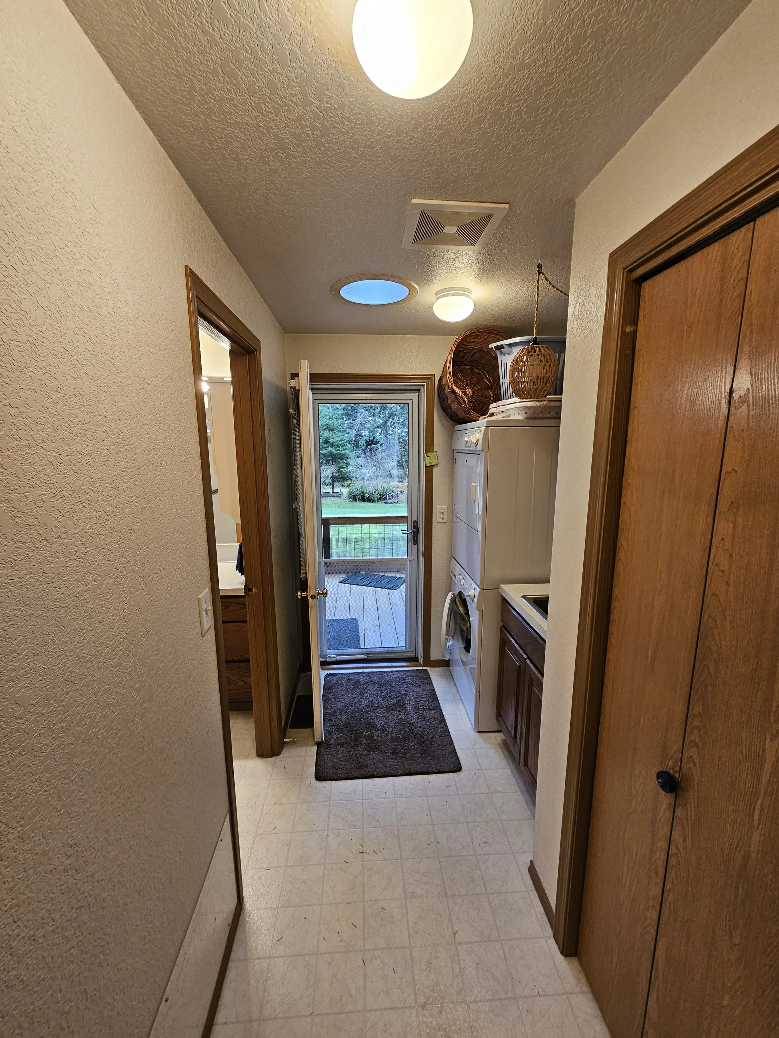 A small laundry area in a home, with a stacked washer and dryer, a door with a glass window leading outside, and a ceiling light. Wicker baskets and clothing are on top of the machines, and there's a black doormat on the floor.