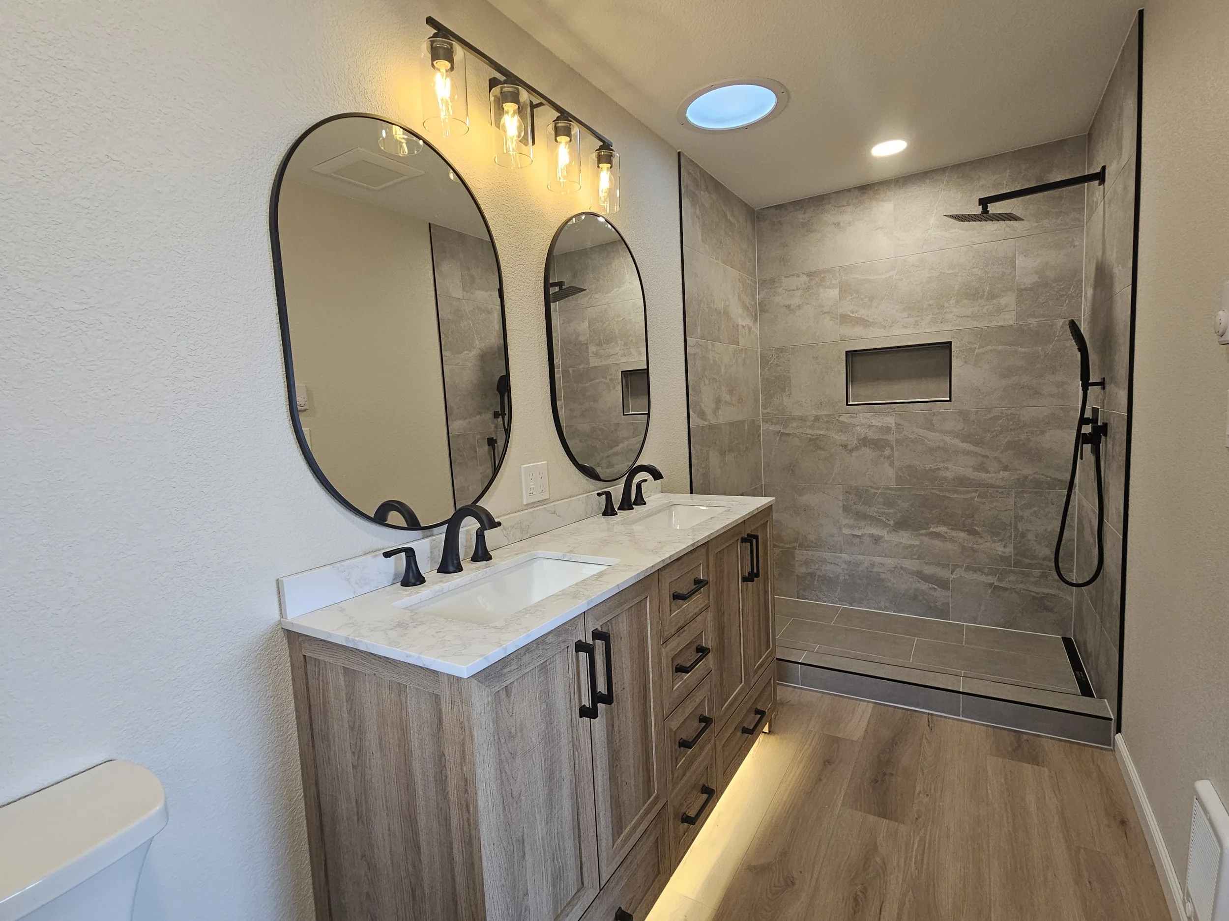A modern bathroom with a double vanity, two oval mirrors, black fixtures, a walk-in shower with gray tile walls and a rainfall showerhead, two ceiling lights, and a skylight.