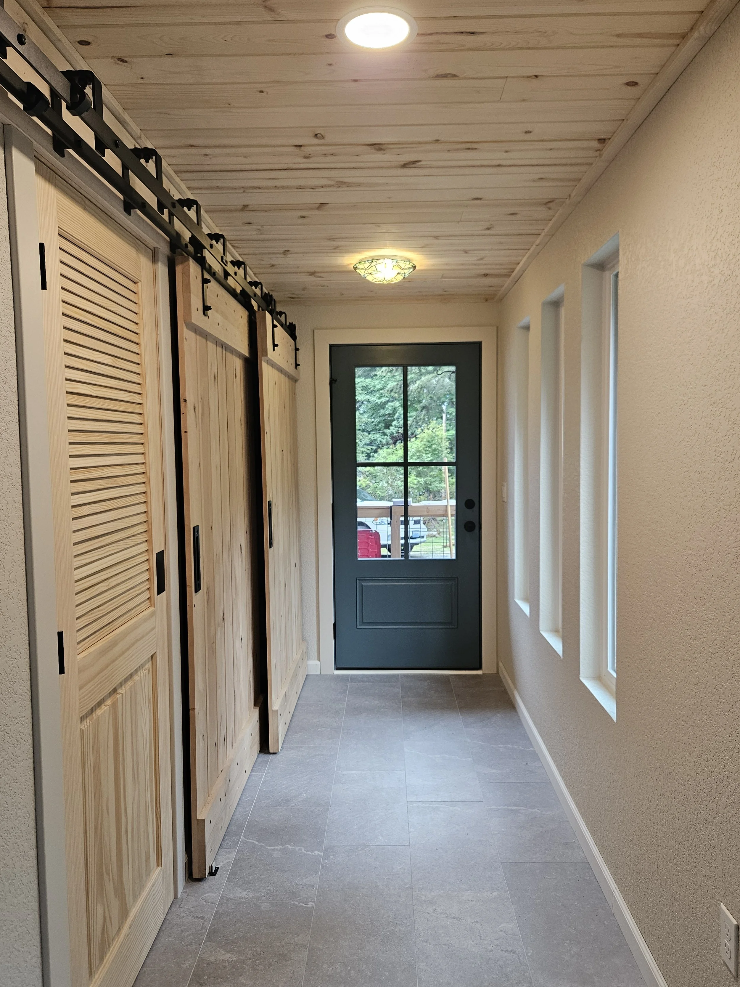 An indoor hallway with a wooden ceiling, three narrow windows on the right wall, a door with a window at the end, and unfinished wooden closet doors on the left side.