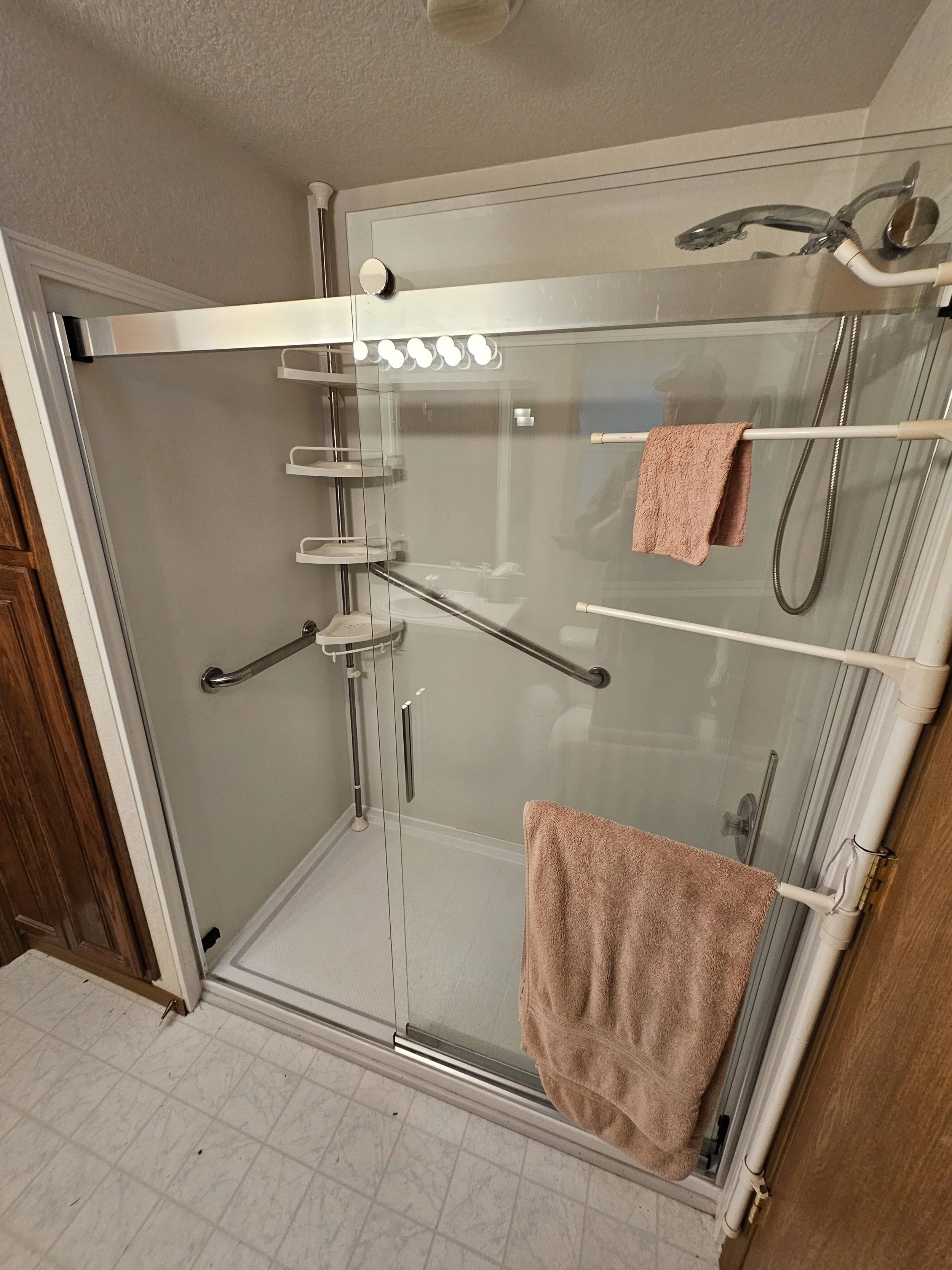 Bathroom shower with glass doors, pink towels, and shower caddy in a corner. The shower has a handrail and a handheld showerhead. The bathroom floor has light-colored tiles.