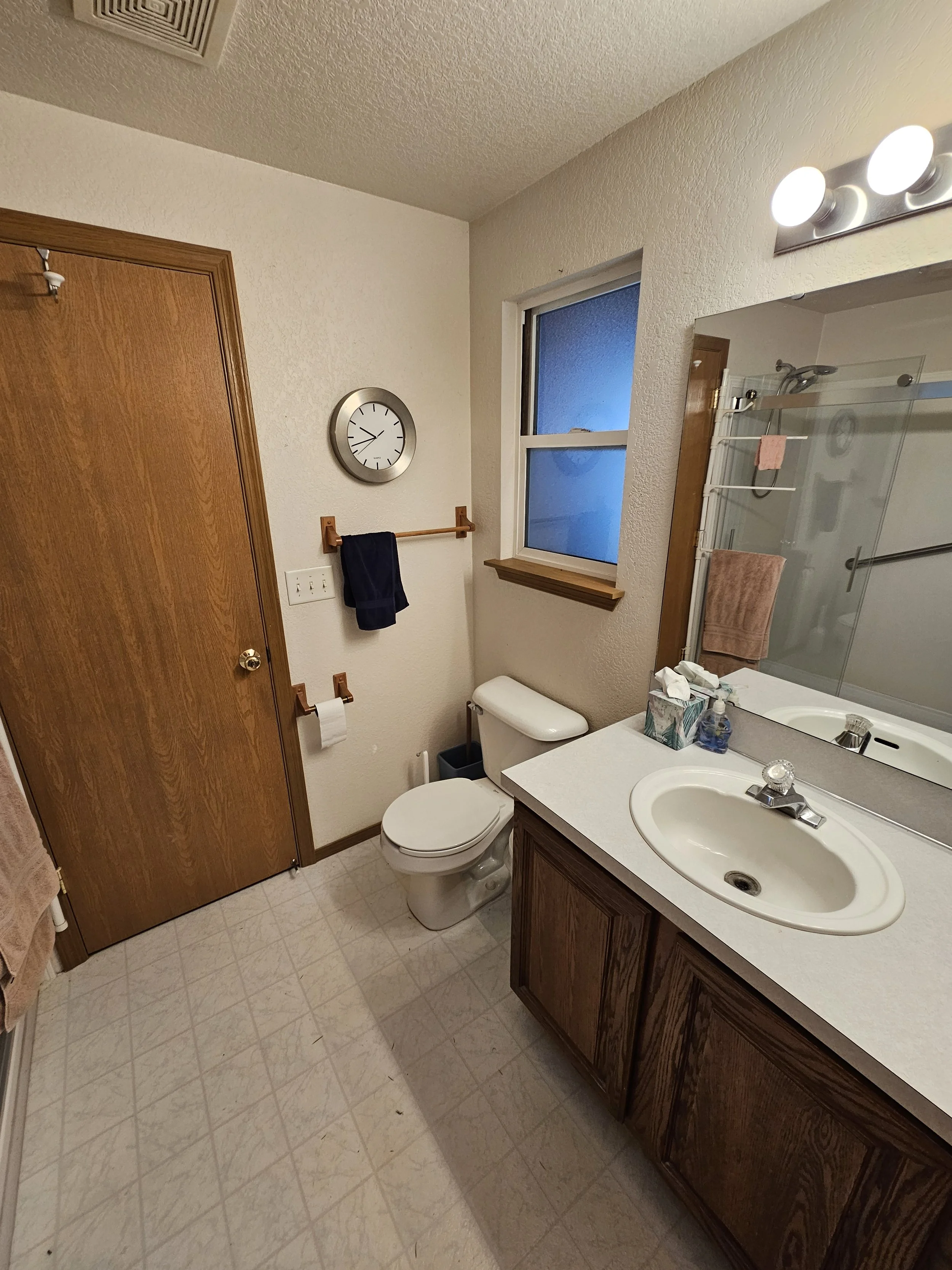 A small bathroom with a wooden door, a round clock on the wall, a window with frosted glass, a bathroom towel rack with one black towel, a toilet, a vanity with a sink, a tissue box, mirror with lights, and a shower with a glass door and pink towels.