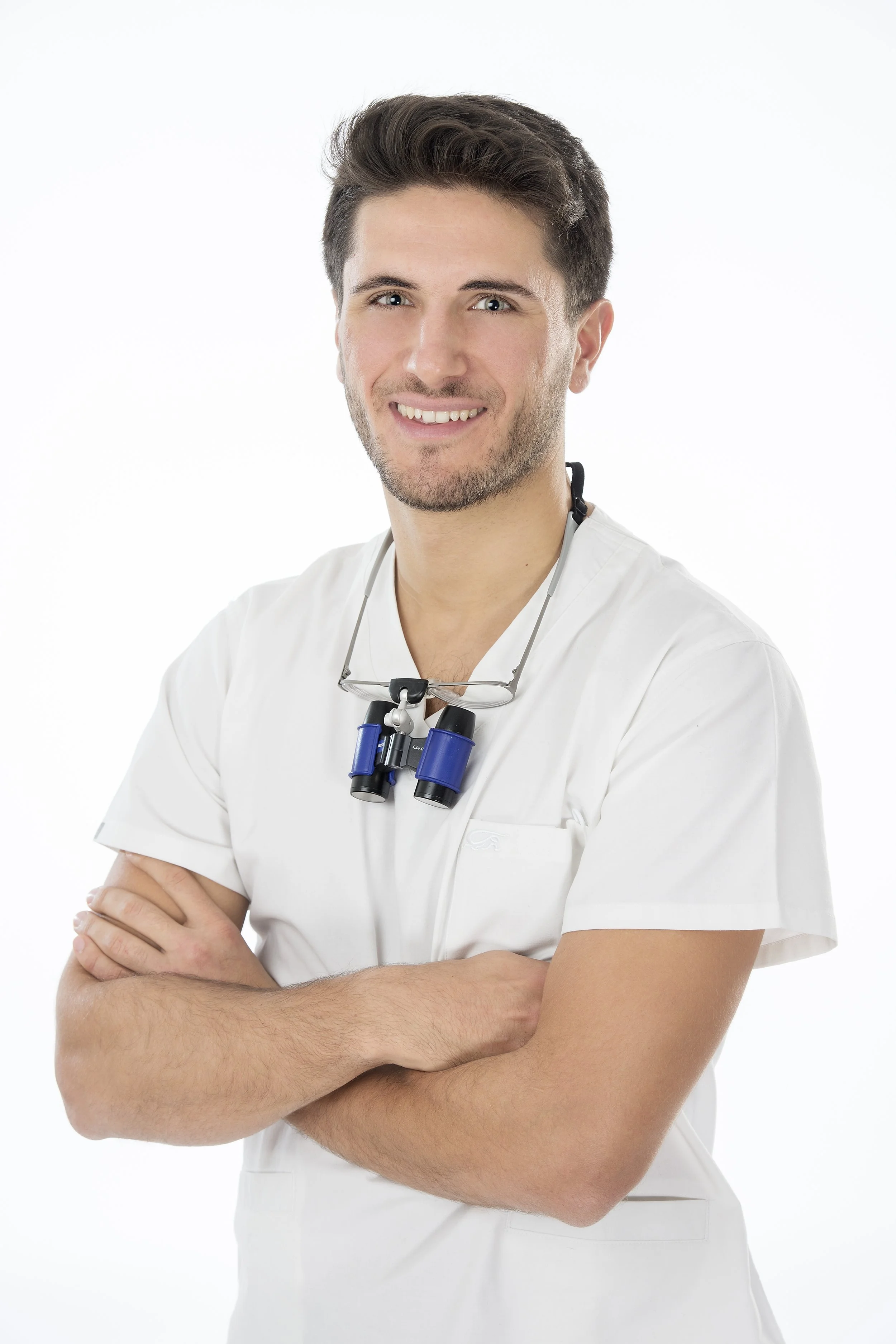 Medico giovane in divisa bianca, con occhiali da ingrandimento appesi al collo, sorride e ha un aspetto empatico.