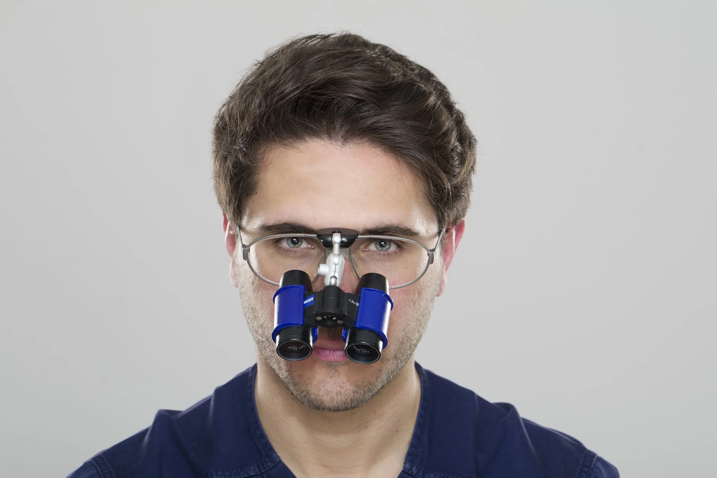 Medico con occhiali da ingrandimento di alta precisione sul naso, guarda direttamente in camera, sfondo grigio.