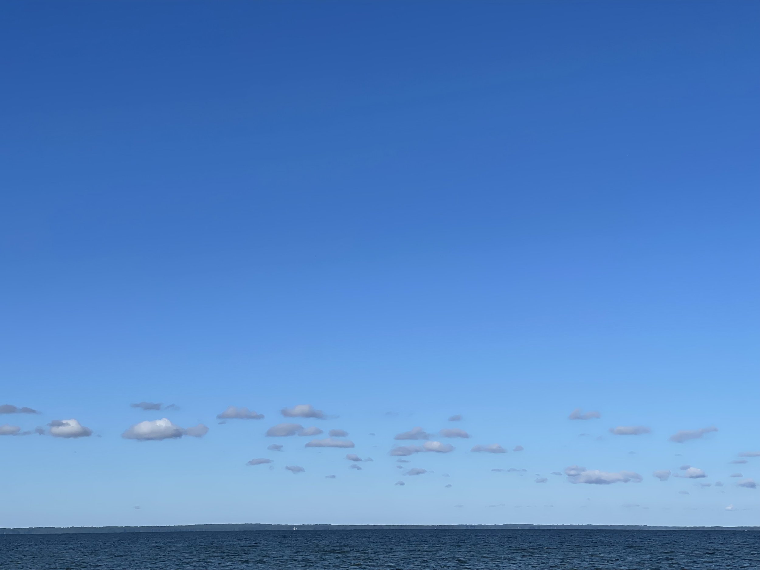 A seascape with calm water, a clear blue sky, and scattered small clouds on the horizon.