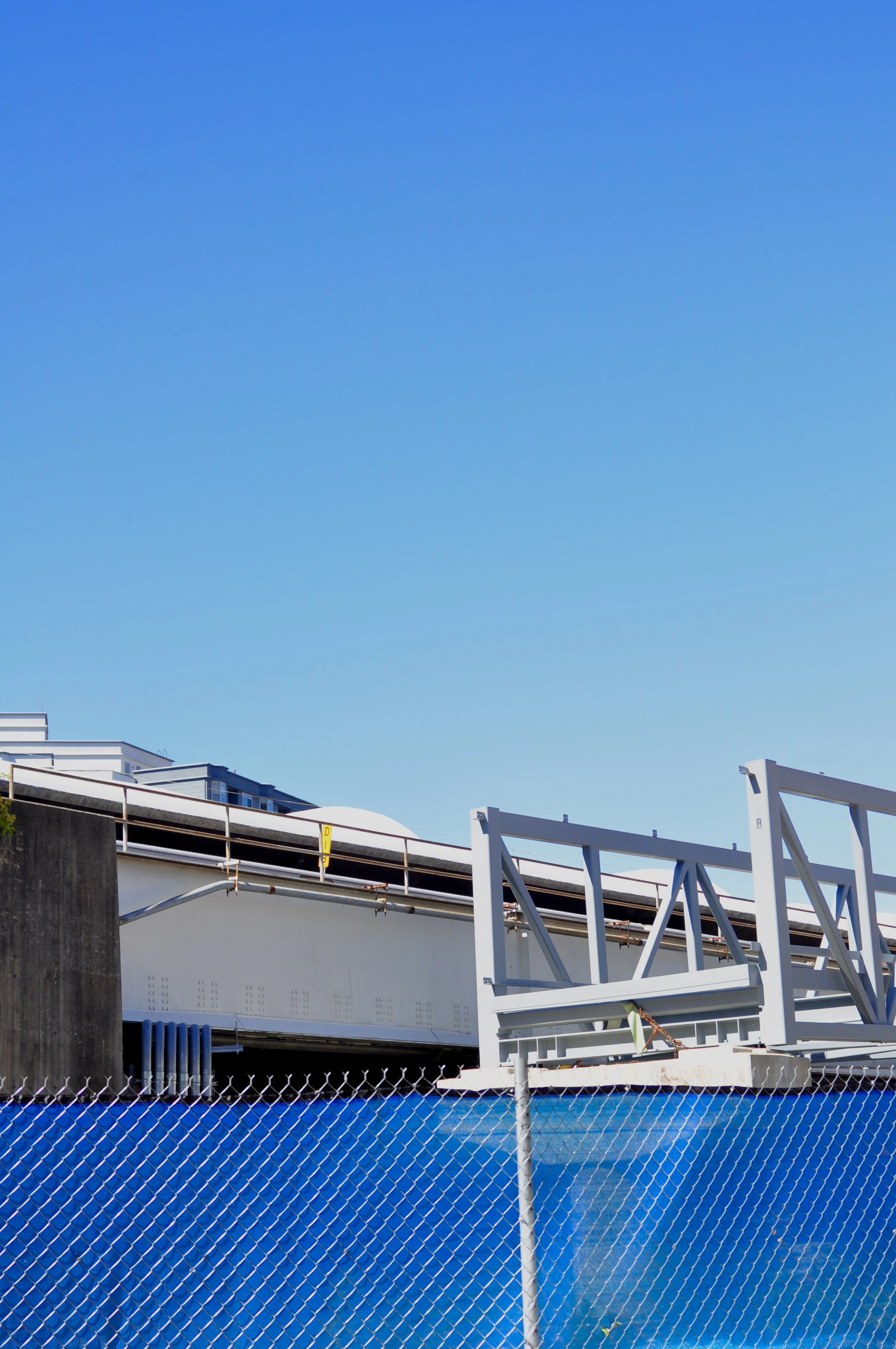 Partial view of a construction site with a blue sky, a white metal structure, a chain-link fence, and a building in the background.