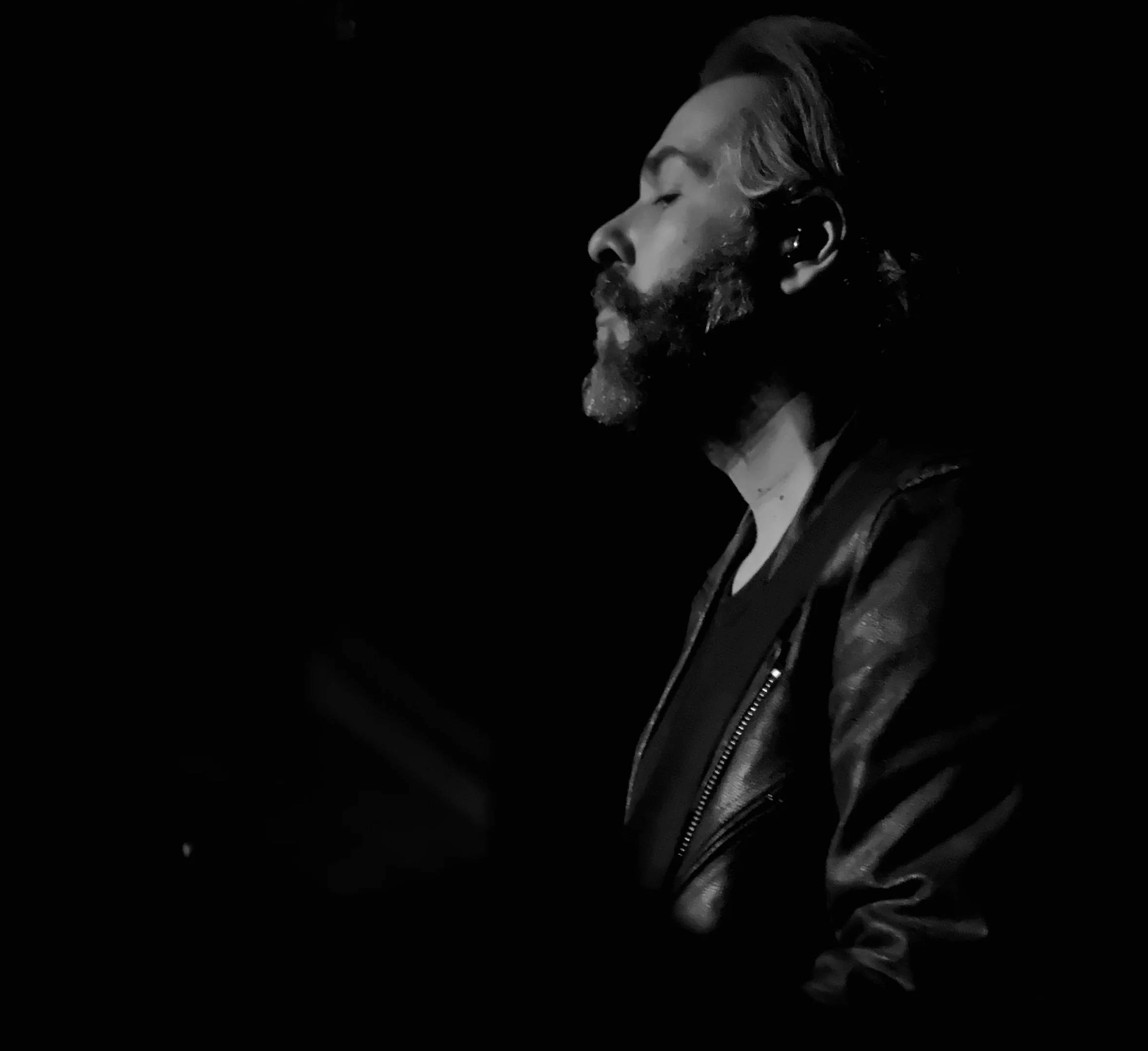 Side profile of a bearded man with closed eyes, wearing a leather jacket and a shirt, against a black background.
