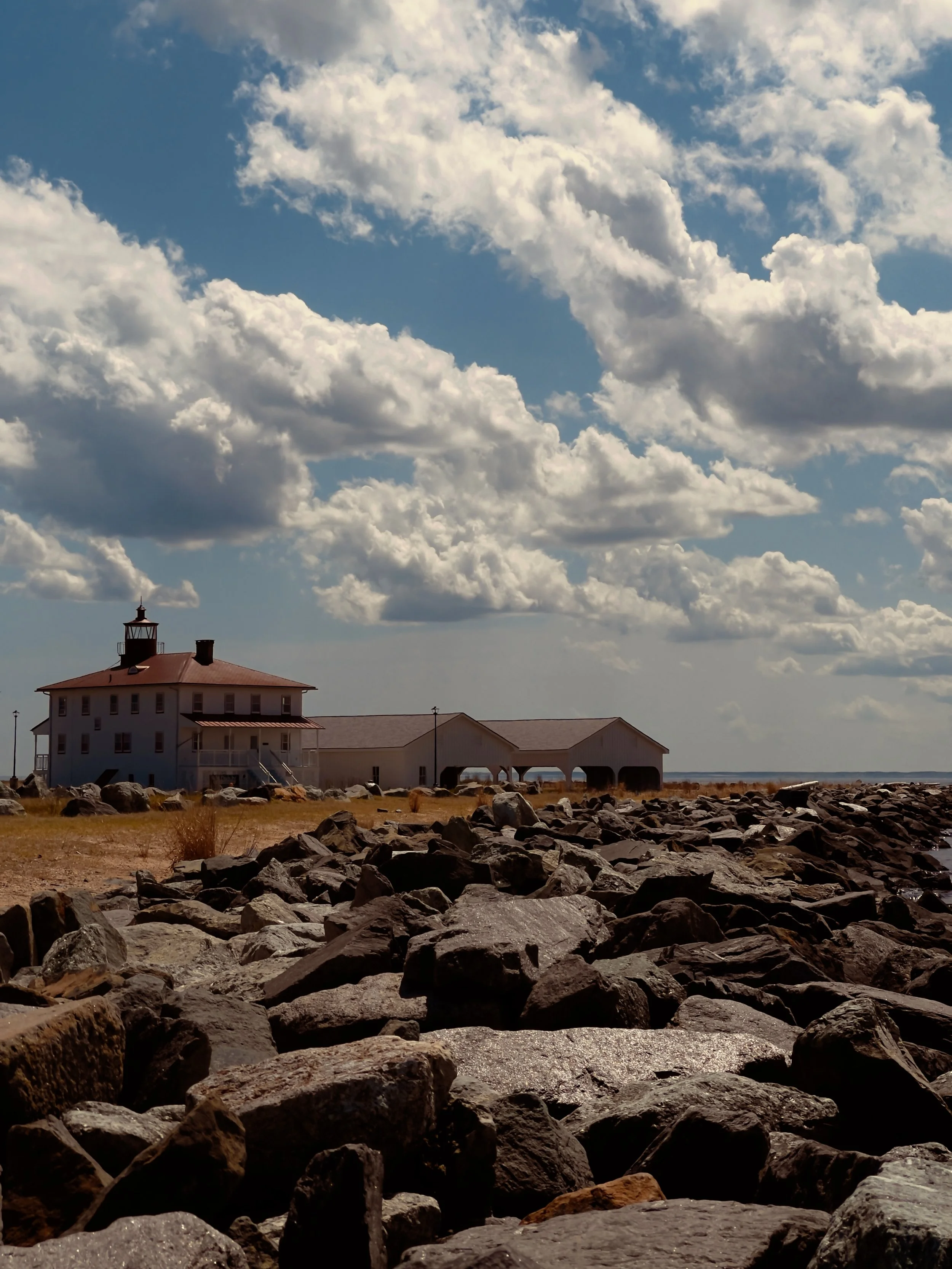 Scenic coastal scene with a rocky shoreline, a lighthouse, and a building under a partly cloudy sky.