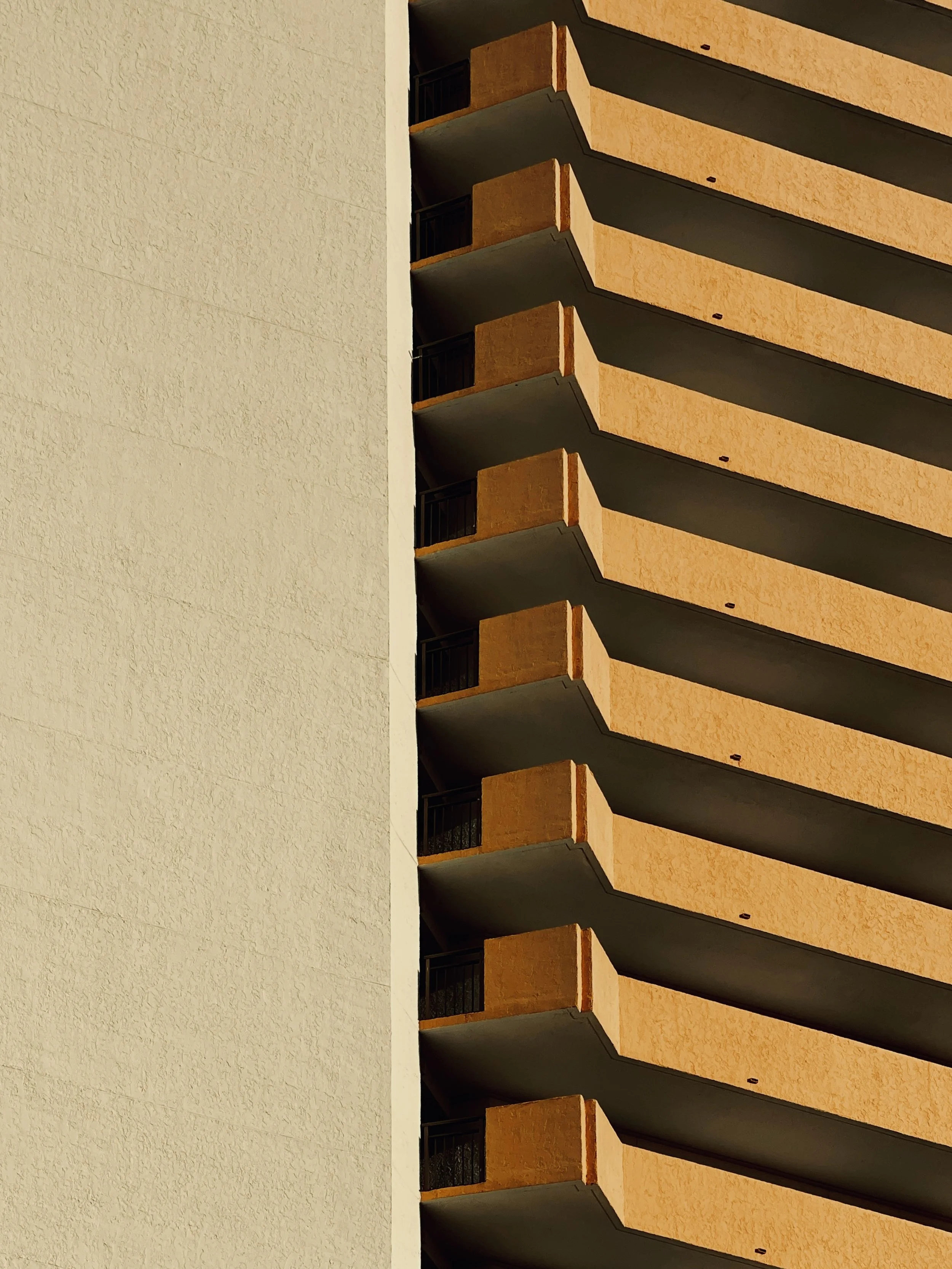 Close-up view of a building with multiple balconies, with one side showing a plain light-colored wall and the other side showing orange balconies with black railings.