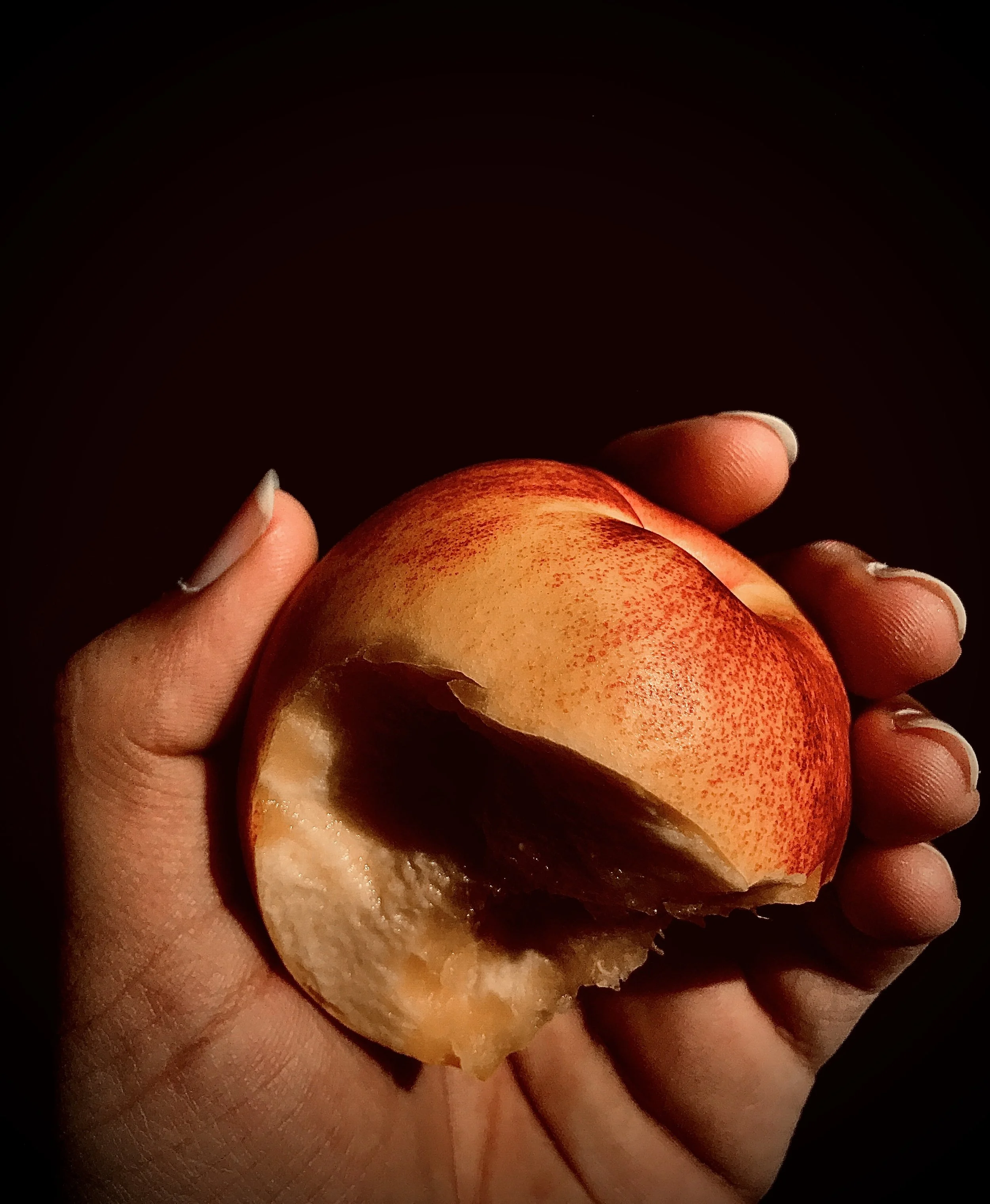A hand holding a partially eaten peach with a bite taken out of it, set against a dark background.