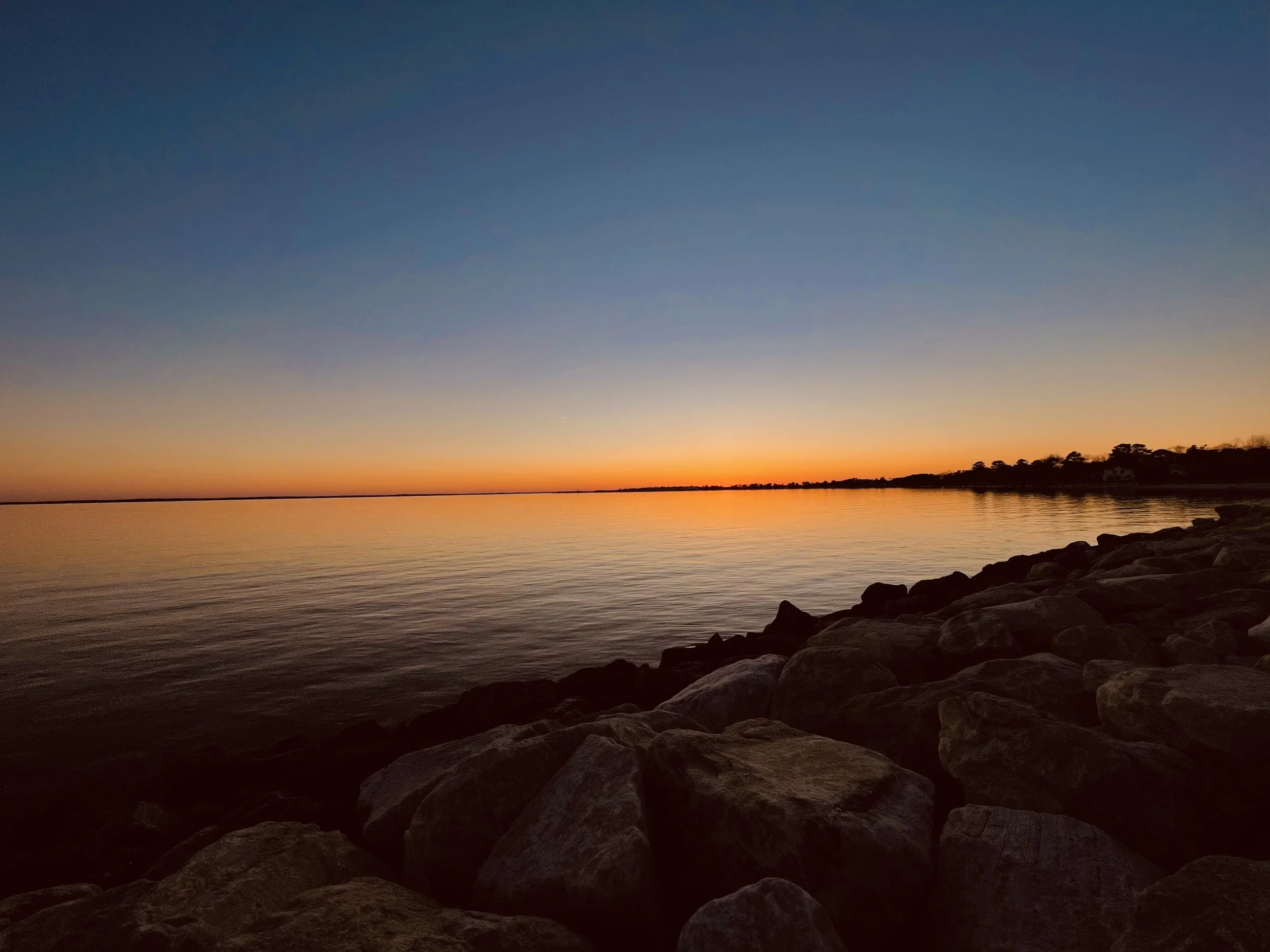 Sunset over a calm body of water with a rocky shoreline and a colorful sky.