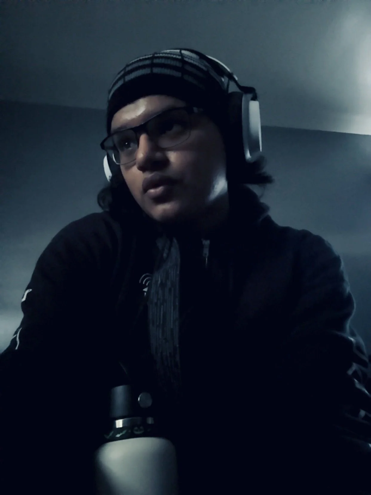 A young person wearing glasses, large headphones, and a striped beanie, looking to the side with a serious expression, in a dimly lit room.