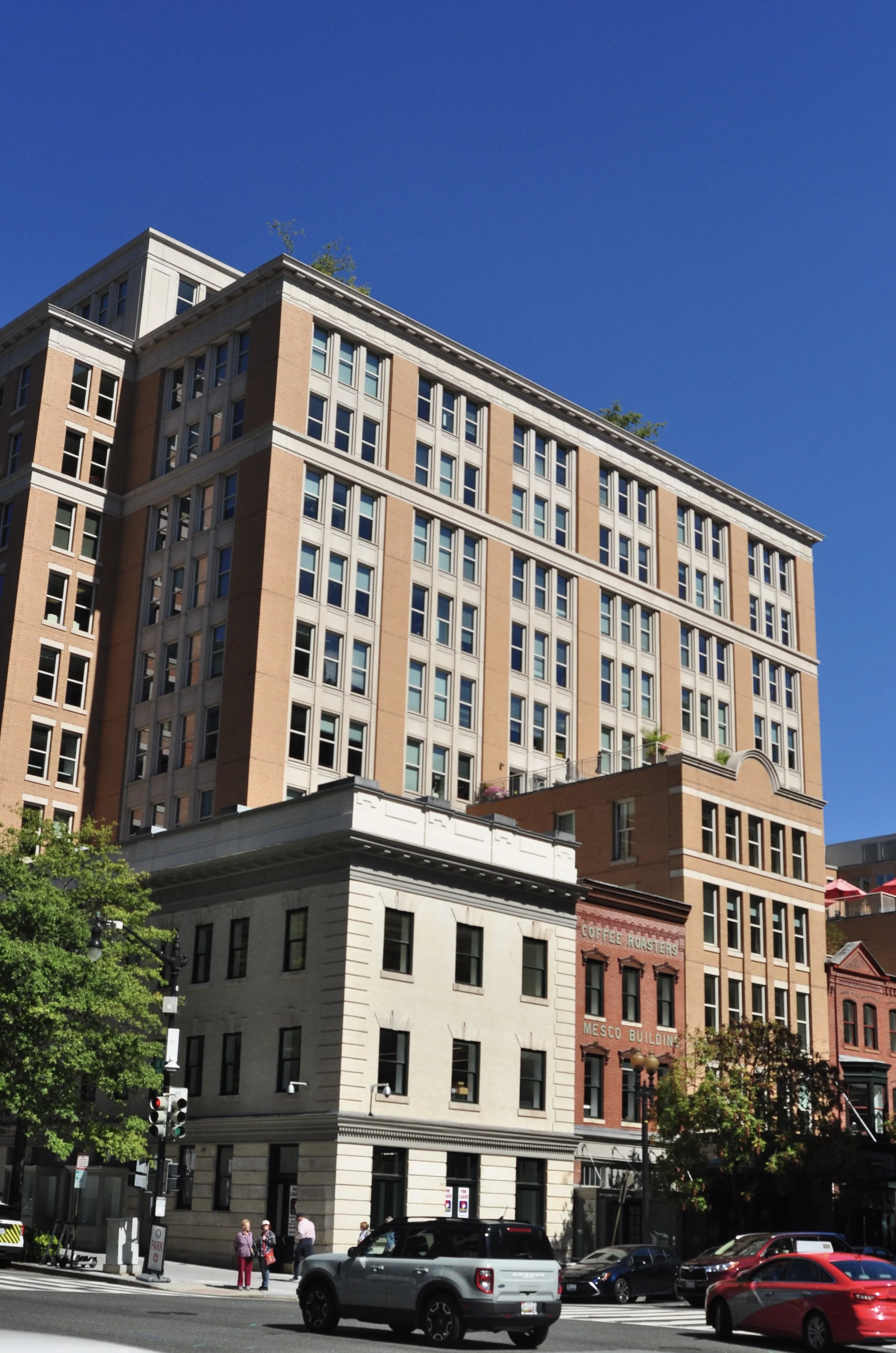 A tall multi-story building with a mix of modern and historic architectural styles, situated on a city street with cars and pedestrians, under a clear blue sky.