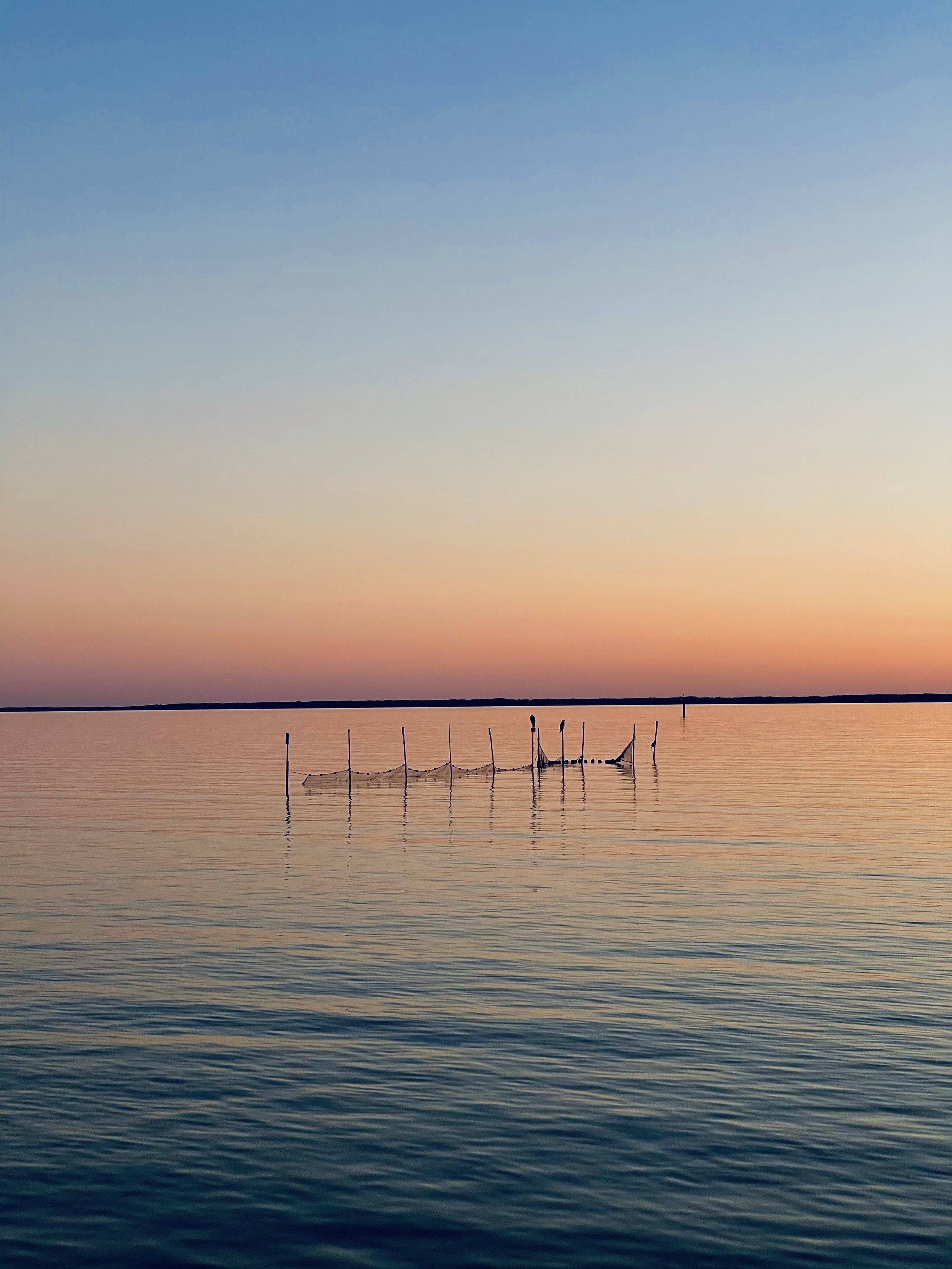 Calm water with a partially submerged boat or structure, reflections on the surface, and a colorful sunset or sunrise sky with shades of pink, orange, and blue.