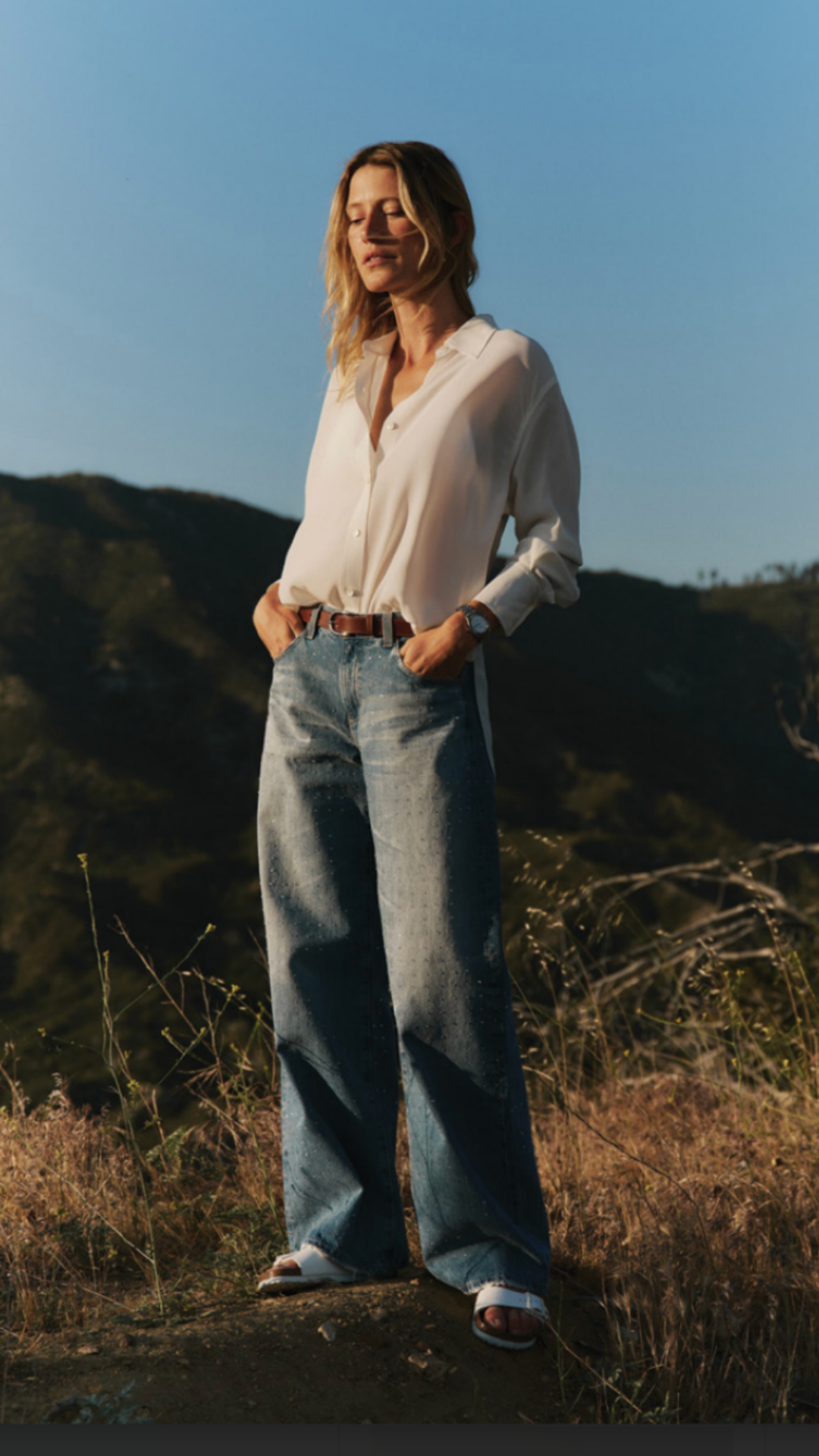 A woman standing outdoors in a natural setting during sunset, wearing a white blouse, jeans, and sandals, with her hands in her pockets and eyes closed.