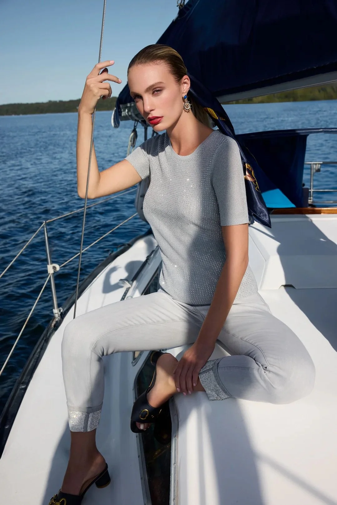 A woman with blonde hair and red lipstick sitting on a yacht, dressed in a silver top, white pants with silver cuffs, and black heels, holding a rope with water in the background.