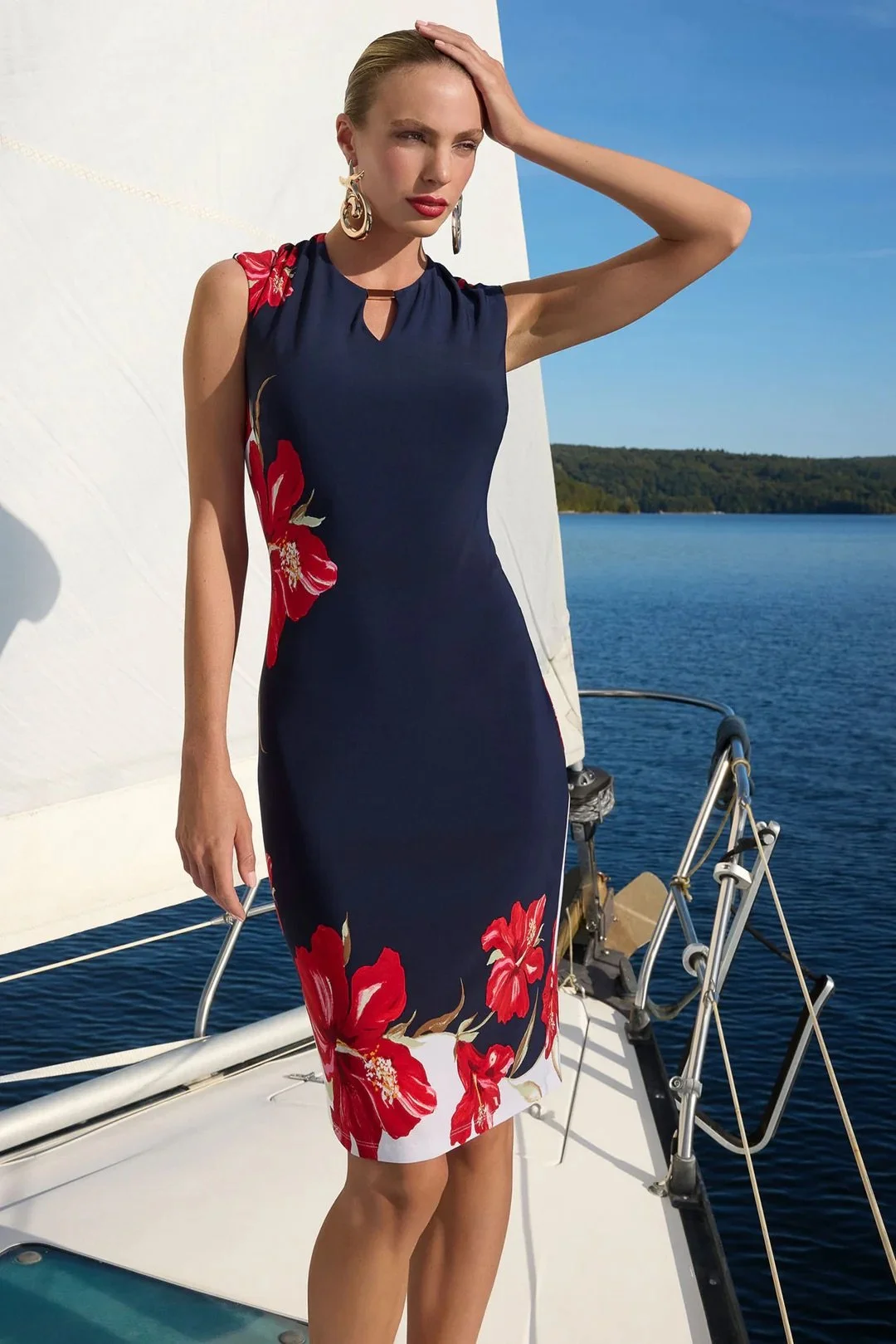 A woman by the water wearing a sleeveless navy dress with large red floral patterns.