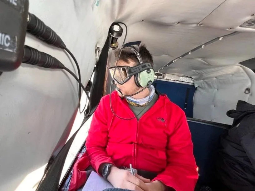 Person wearing headphones, safety glasses, and a red jacket, taking notes inside an aircraft with a window showing the sky outside.