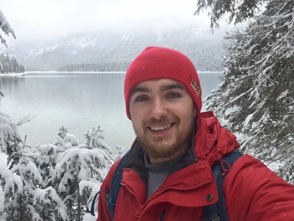 A man wearing a red beanie and red jacket taking a selfie in a snowy forest near a lake with snow-covered trees and mountains in the background.