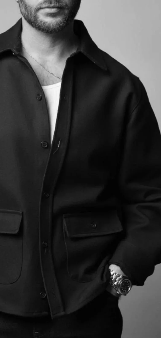 Black and white photo of a man wearing a black jacket over a white T-shirt, with a watch on his wrist and a necklace, standing against a plain background. Guillaume Boucher