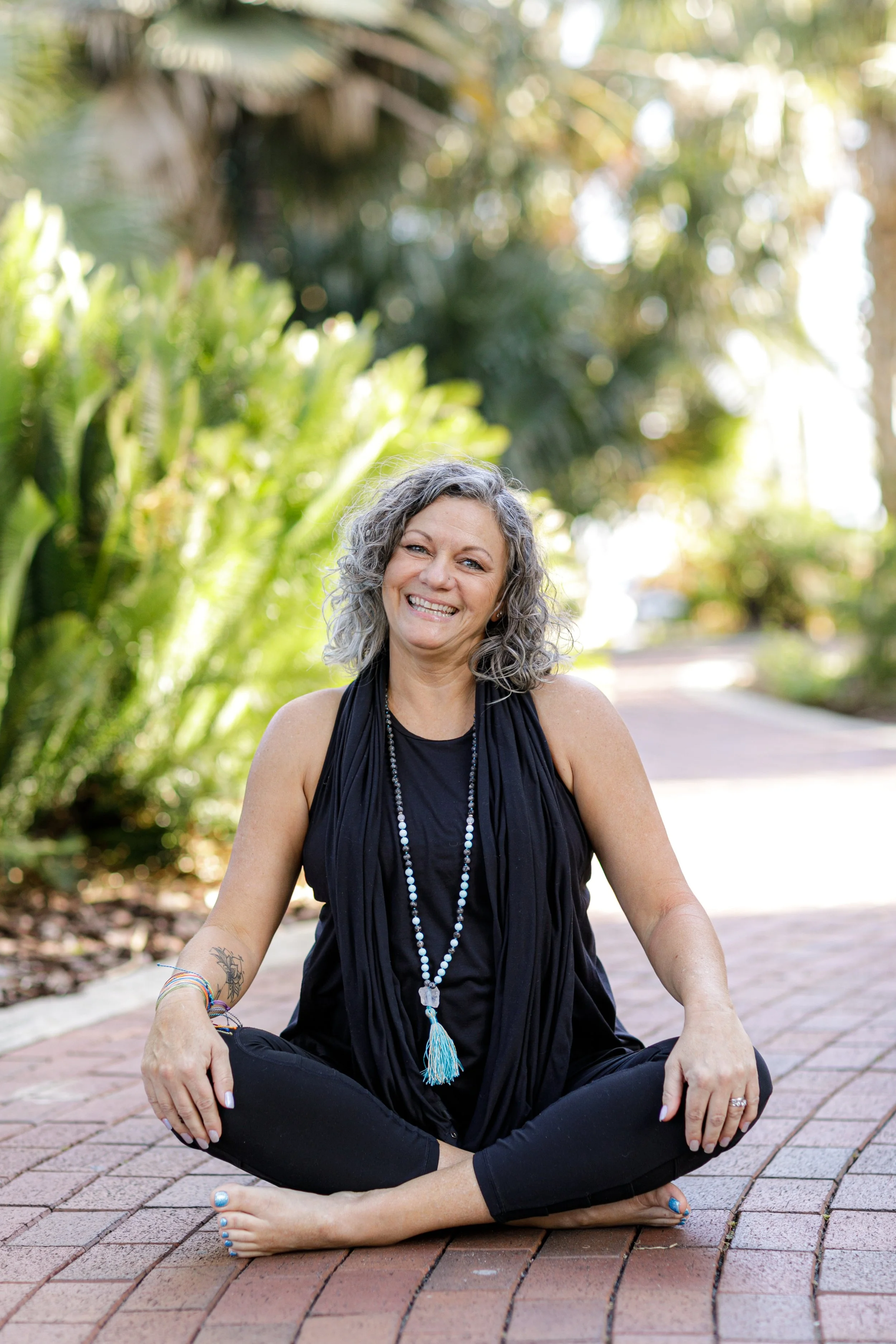 Smile and relaxed woman sitting cross-legged outdoors on brick path, with lush green plants and trees in background.