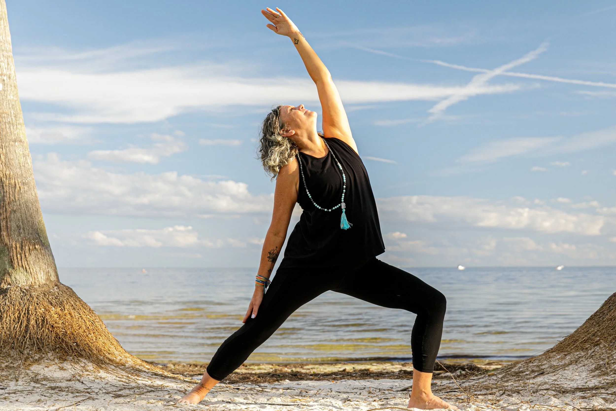 Woman practicing yoga outdoors on the beach, standing in a wide stance, reaching one arm upward, with a scenic ocean and sky background.