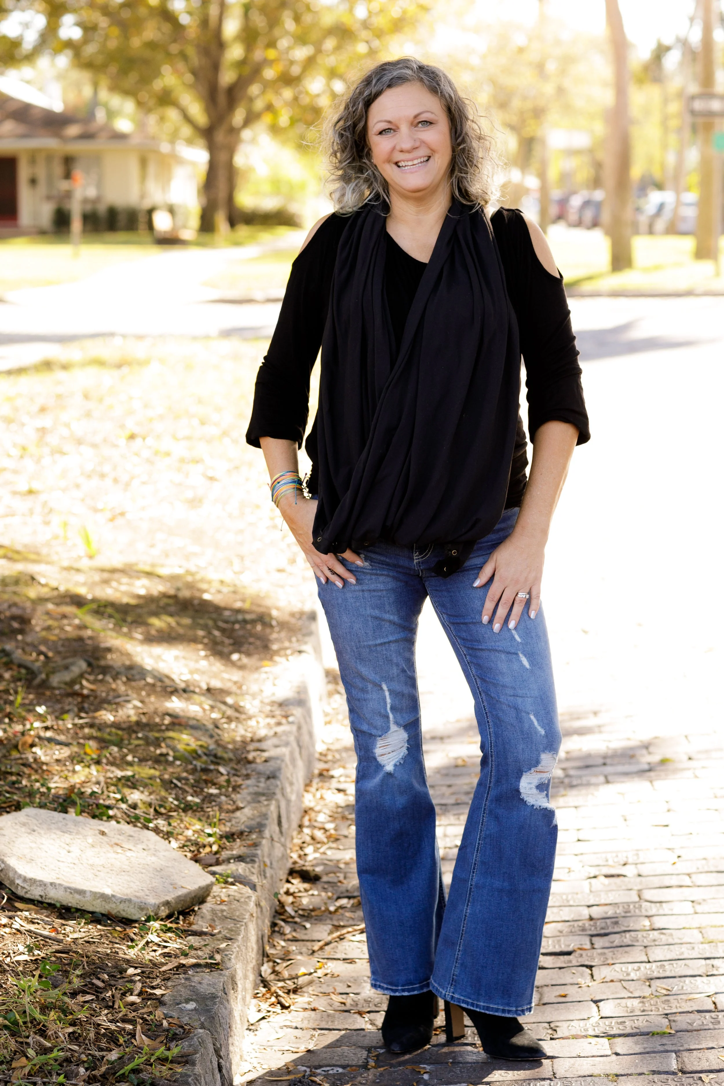 Woman with curly gray hair wearing a black top and ripped jeans, standing outdoors on a sidewalk with trees and houses in the background, smiling.