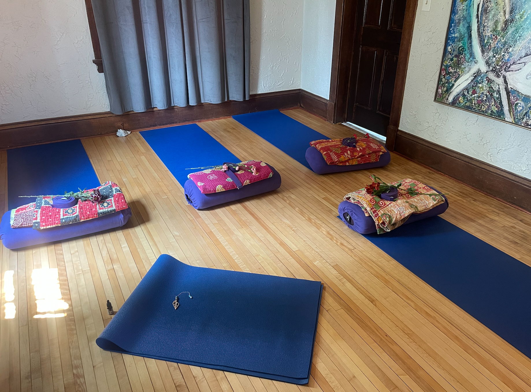 A yoga studio with four blue yoga mats aligned on a wooden floor, each with a decorated bolster and a flower. One blue yoga mat is in the foreground, slightly tilted, with a small bottle and a key on it.