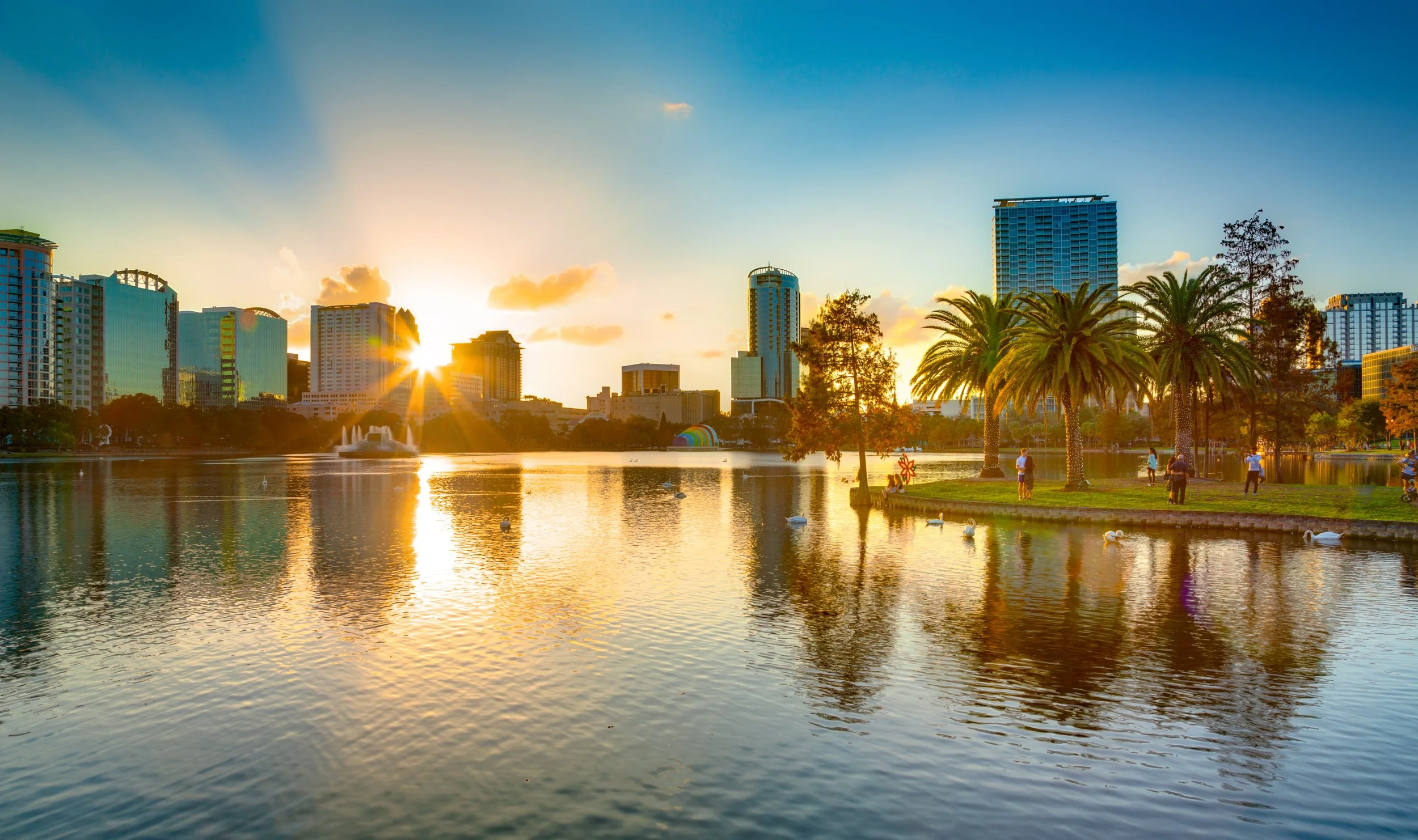 Sunset over a city skyline with high-rise buildings, a lake with swan-shaped paddle boats, palm trees, and people by the water
