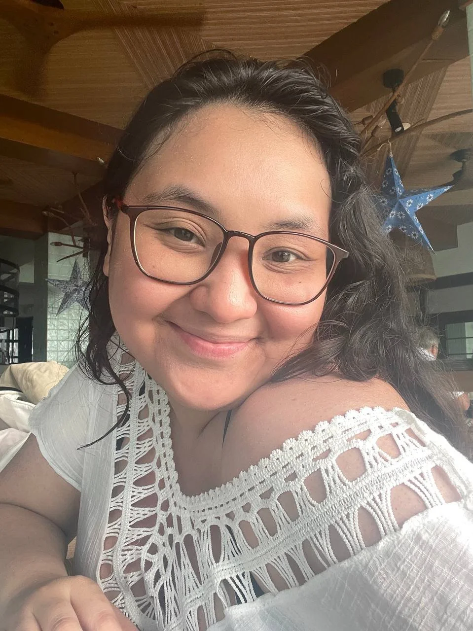 Close-up of a smiling woman with glasses, dark wavy hair, wearing a white crochet top, indoors with wooden ceiling and hanging blue star decoration.