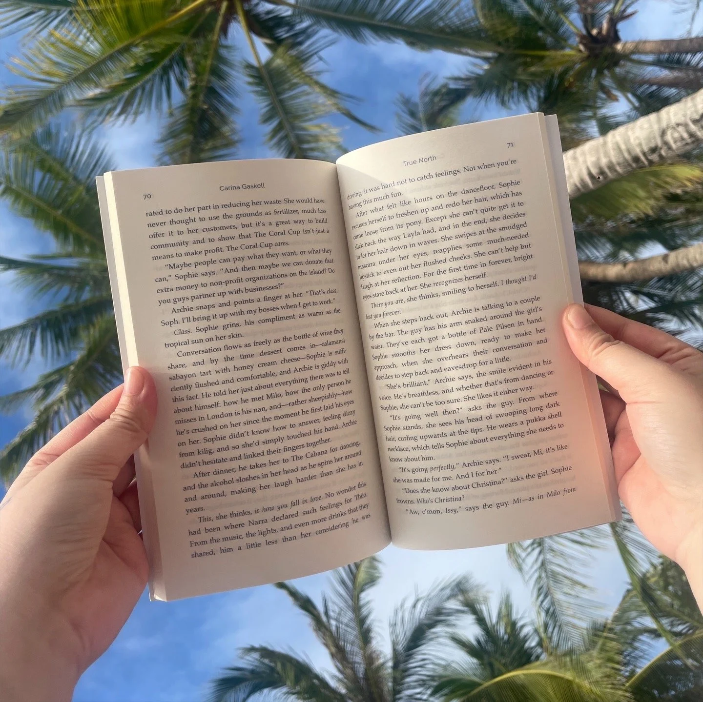 A person holding an open book outdoors, with palm trees and a blue sky in the background.