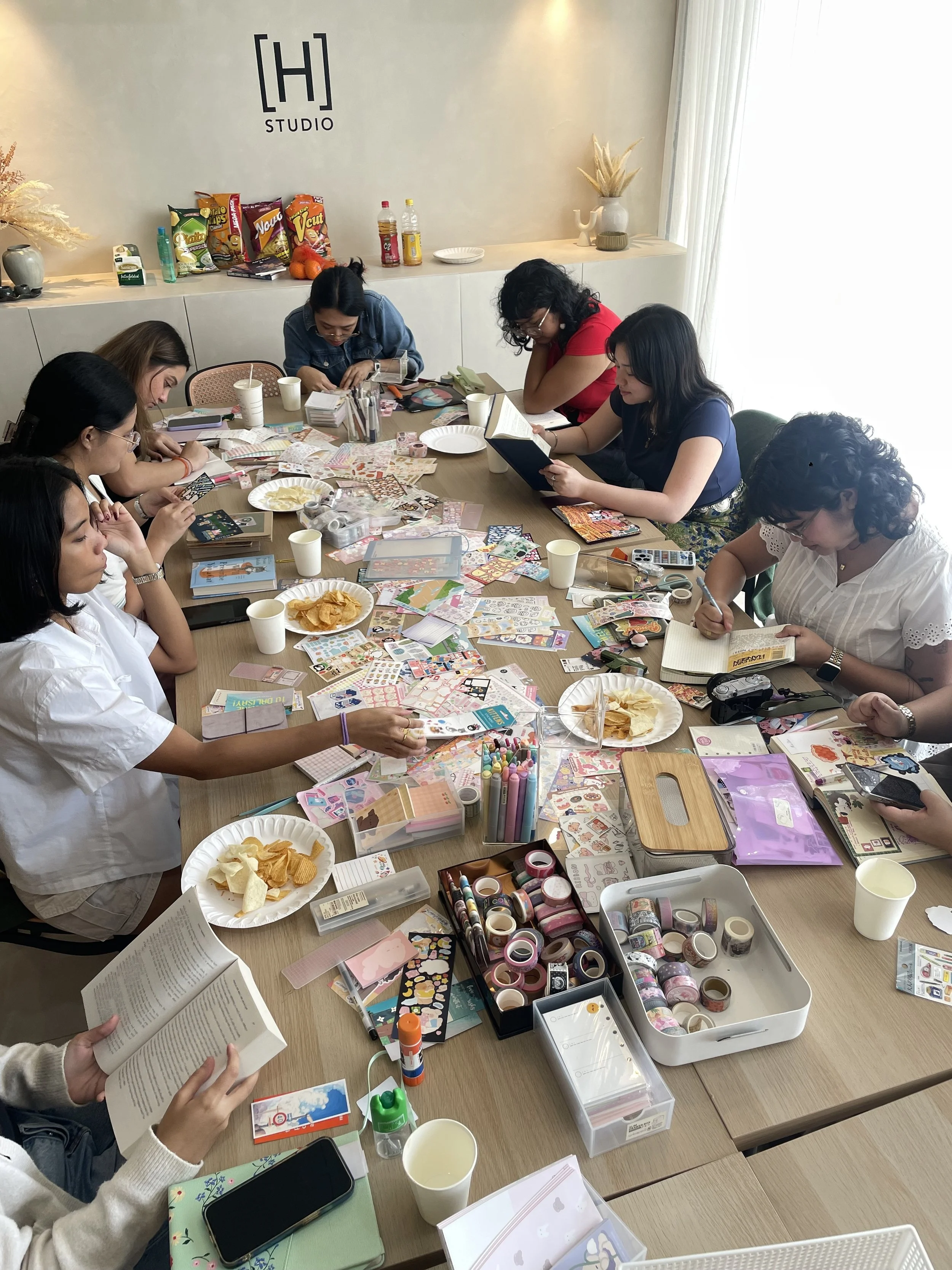 A group of women sitting around a cluttered table covered with stickers, washi tapes, notebooks, and snacks, engaging in arts and crafts activities in a well-lit room with a sign that reads 'H Studio' on the wall.