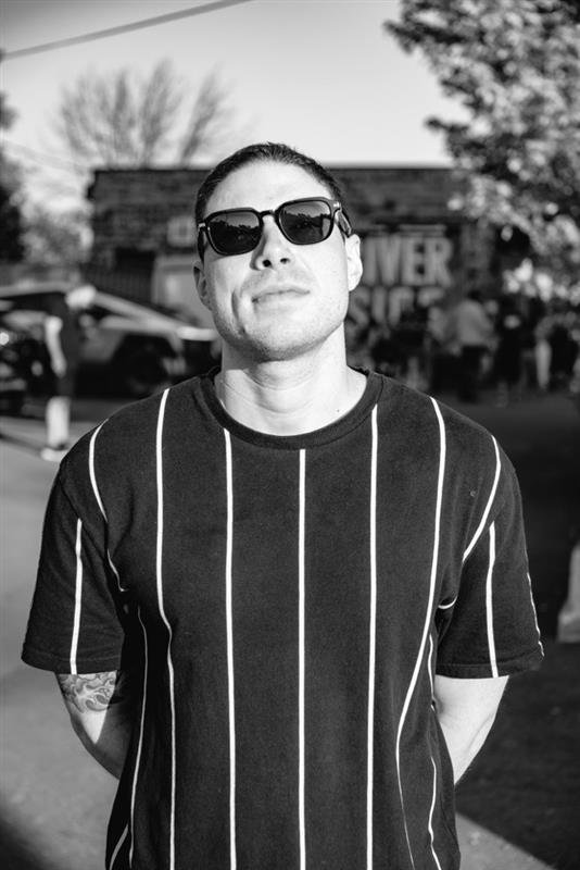 An executive producer wearing sunglasses and a black t-shirt with white stripes standing outdoors on a sunny day.