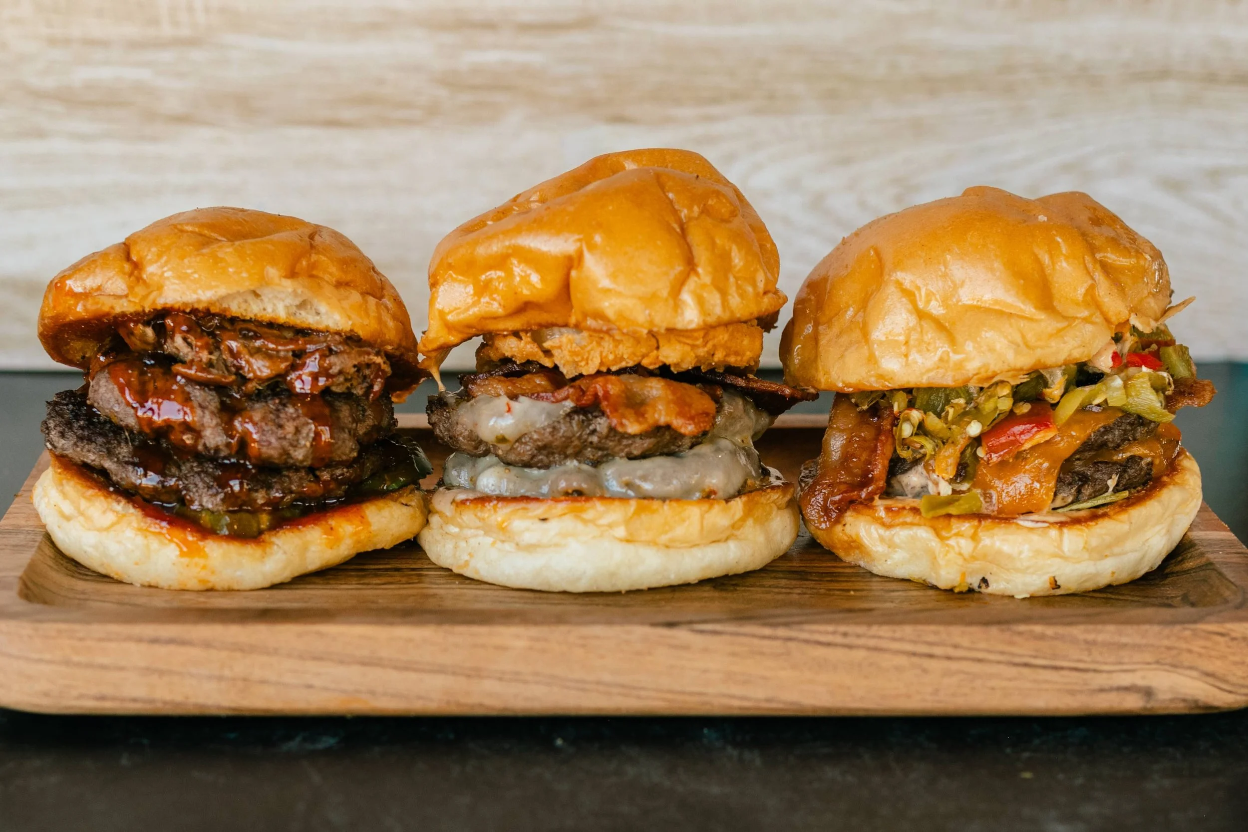 Three different burgers with a variety of toppings on a wooden serving board.
