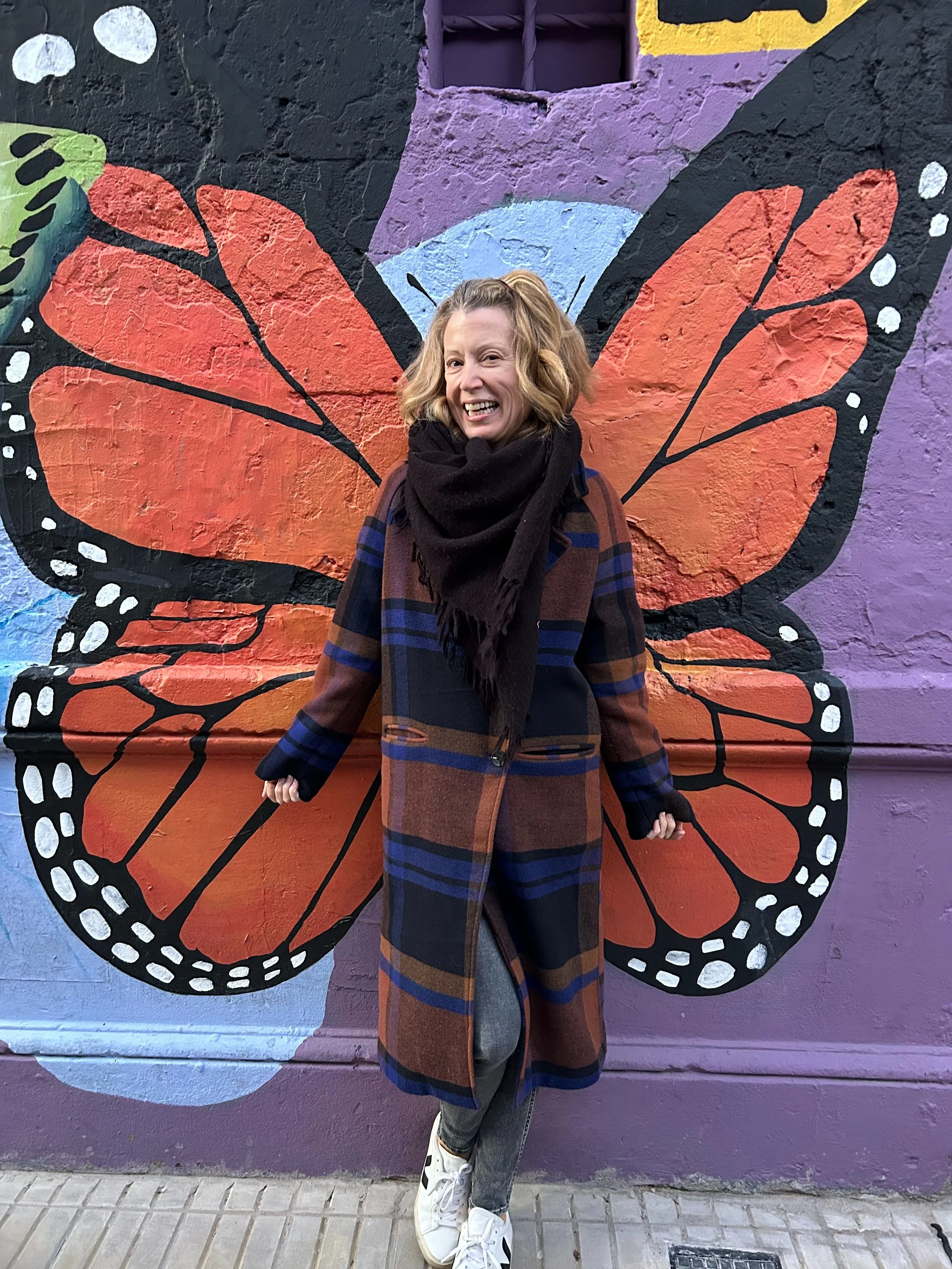 A woman standing in front of a colorful mural of a butterfly painted on a brick wall, with her arms outstretched to resemble part of the butterfly's wings.