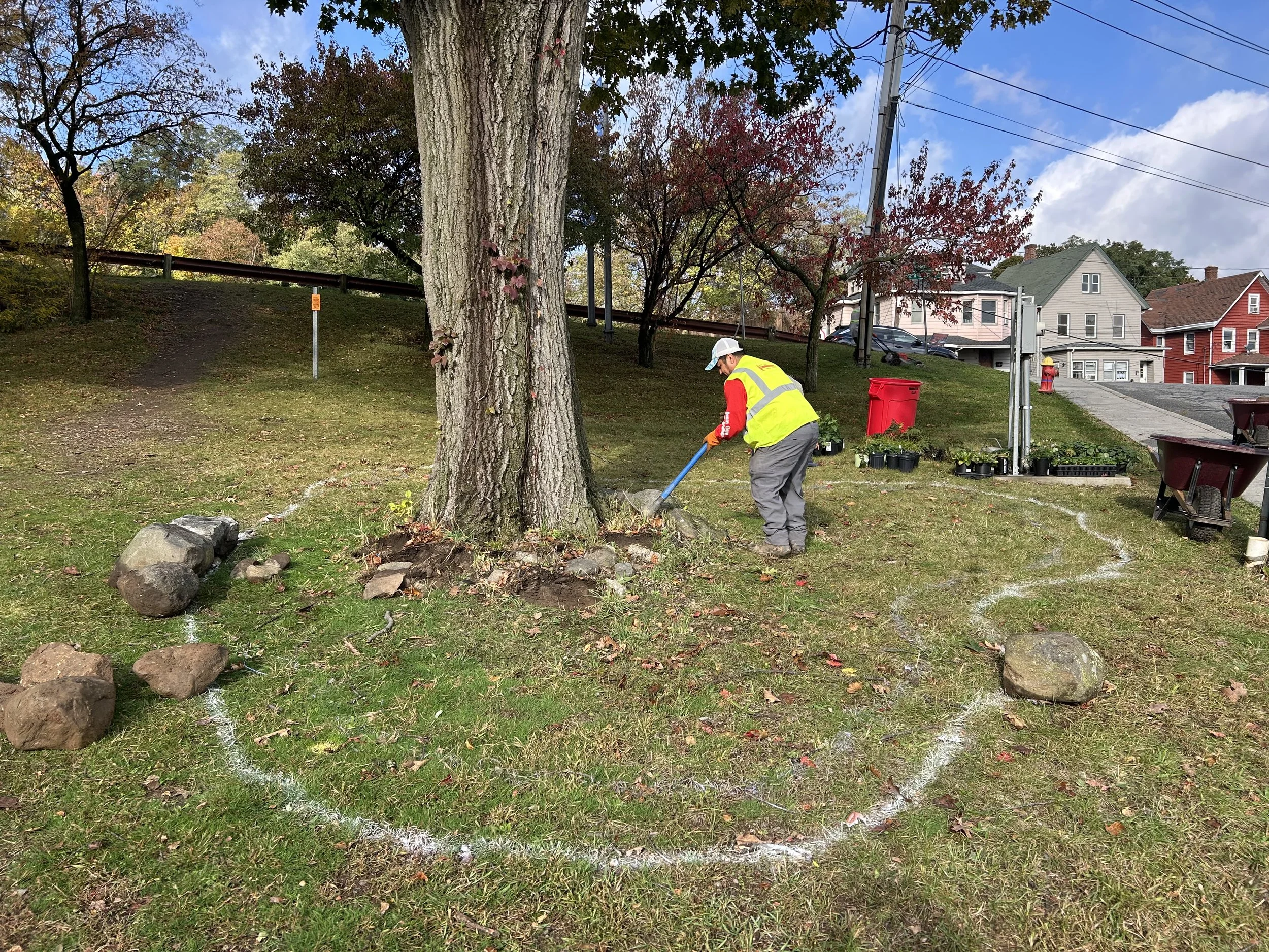 A person wearing a yellow safety vest and gray pants is planting or tending to plants near a large tree in a suburban neighborhood. There are houses, power lines, and a red trash bin in the background, and the sky is partly cloudy.