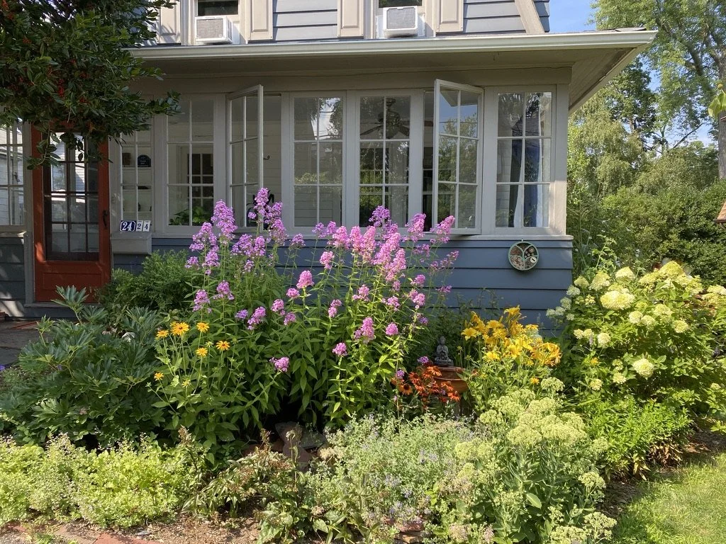 Colorful garden with pink, yellow, and green flowering plants in front of a house with large windows and a red front door.
