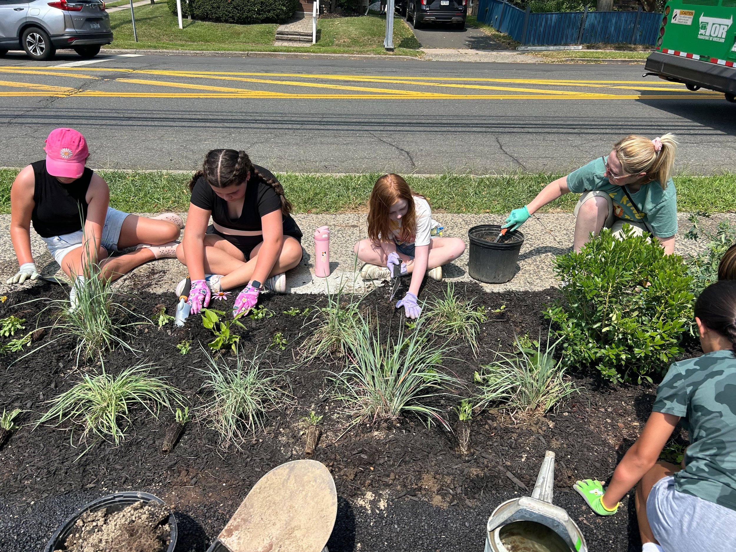 Children planting flowers in a flower bed on a sidewalk, with cars parked on the street behind them.