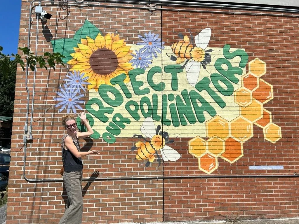 A mural on a brick wall features sunflowers, bees, honeycombs, and the phrase "Protect Our Pollinators" in green letters. A woman in sunglasses is pointing at the mural.