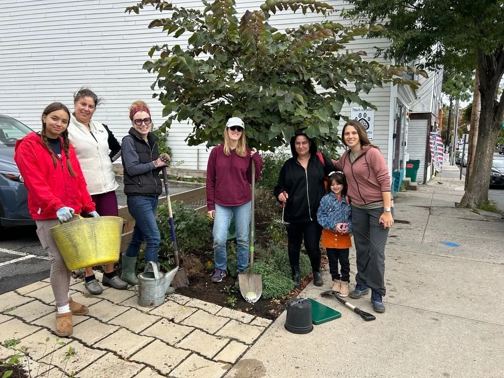 Group of seven women and one young girl planting flowers and doing yard work outdoors on a sidewalk, with a white building and trees in the background.