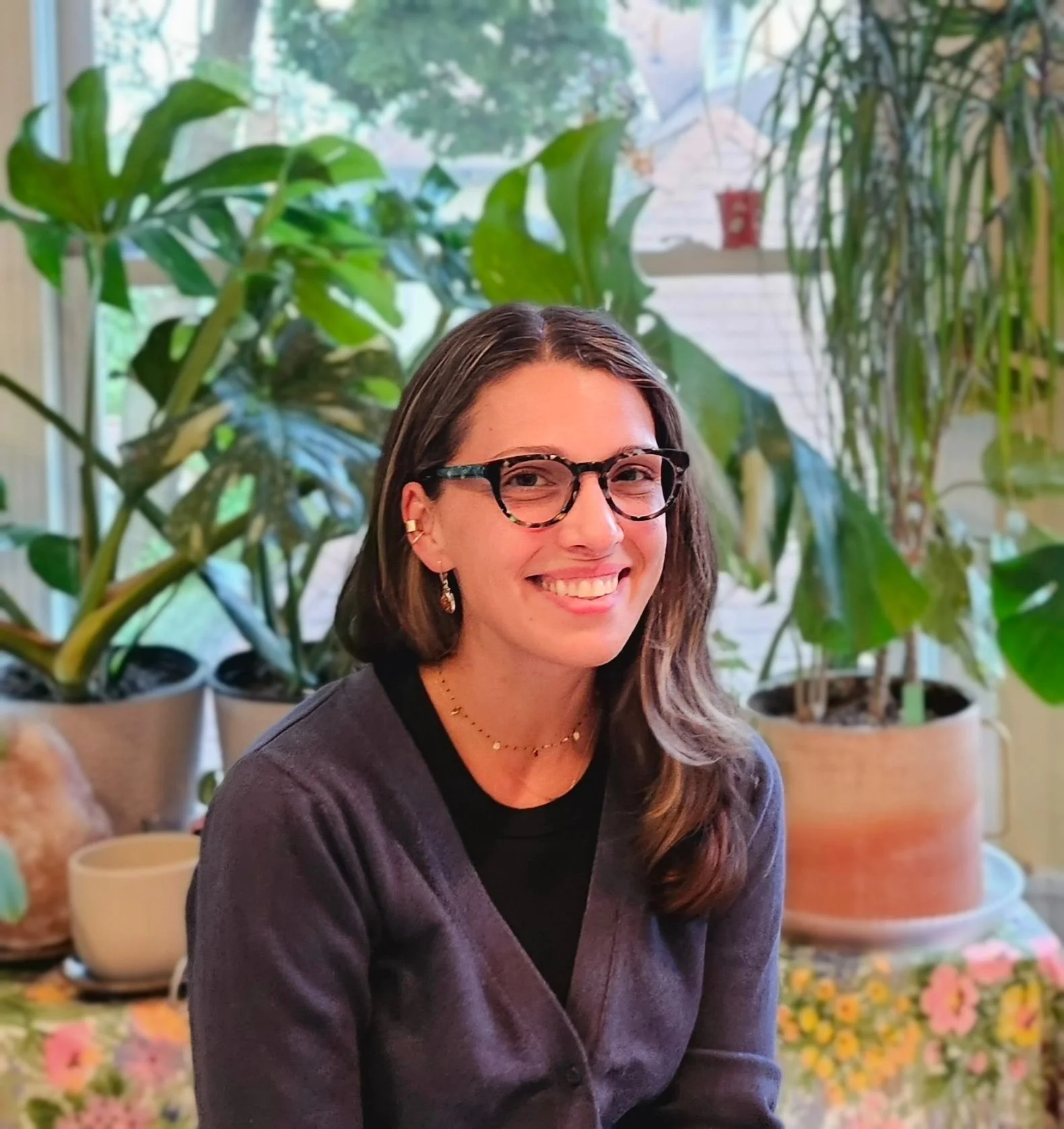 A woman with shoulder-length brown hair, glasses, and earrings smiling indoors with large green houseplants and potted flowers in the background.