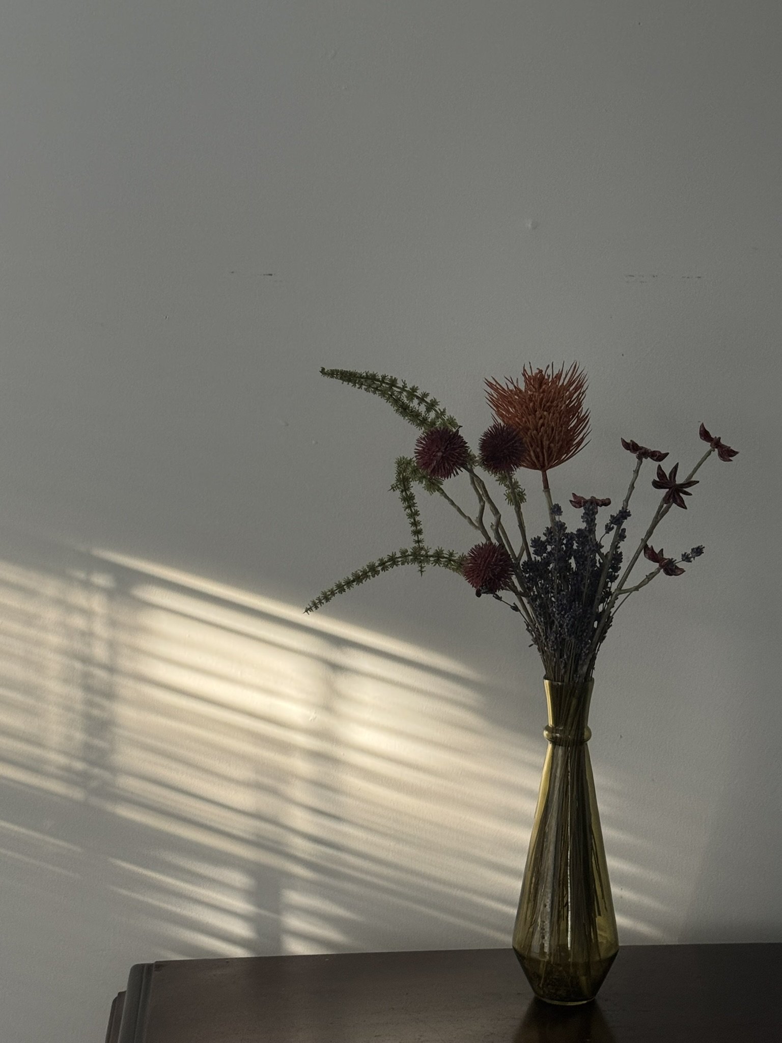 A brown glass vase with dried flowers, including green, orange, and purple blossoms, sitting on a dark wooden surface, with sunlight creating shadows on the white wall behind.