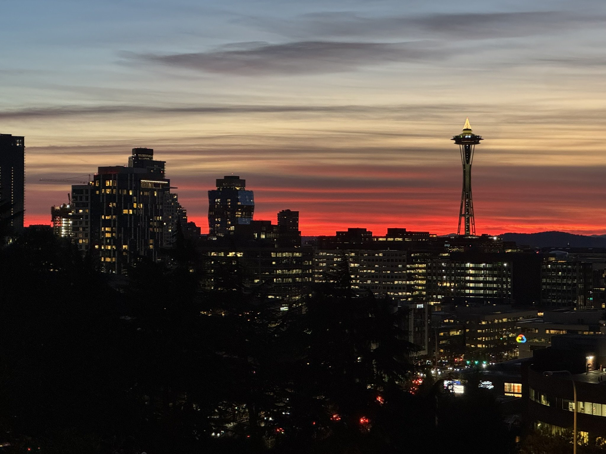 City skyline at sunset with the Space Needle, illuminated buildings, and colorful sky with orange, pink, and blue hues.