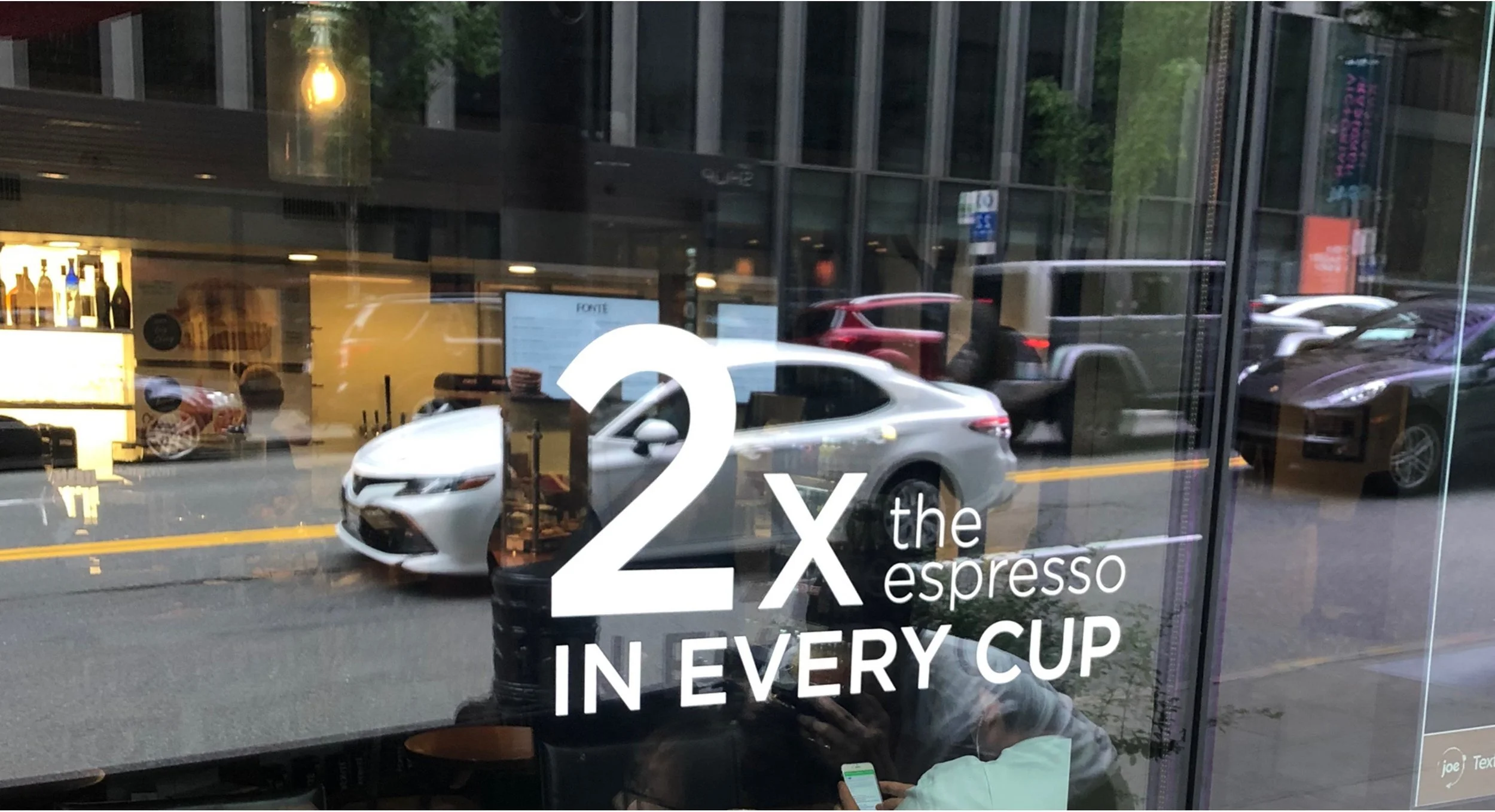Reflection in a cafe window showing cars on the street and text promoting espresso drinks, stating '2 x the espresso in every cup.'