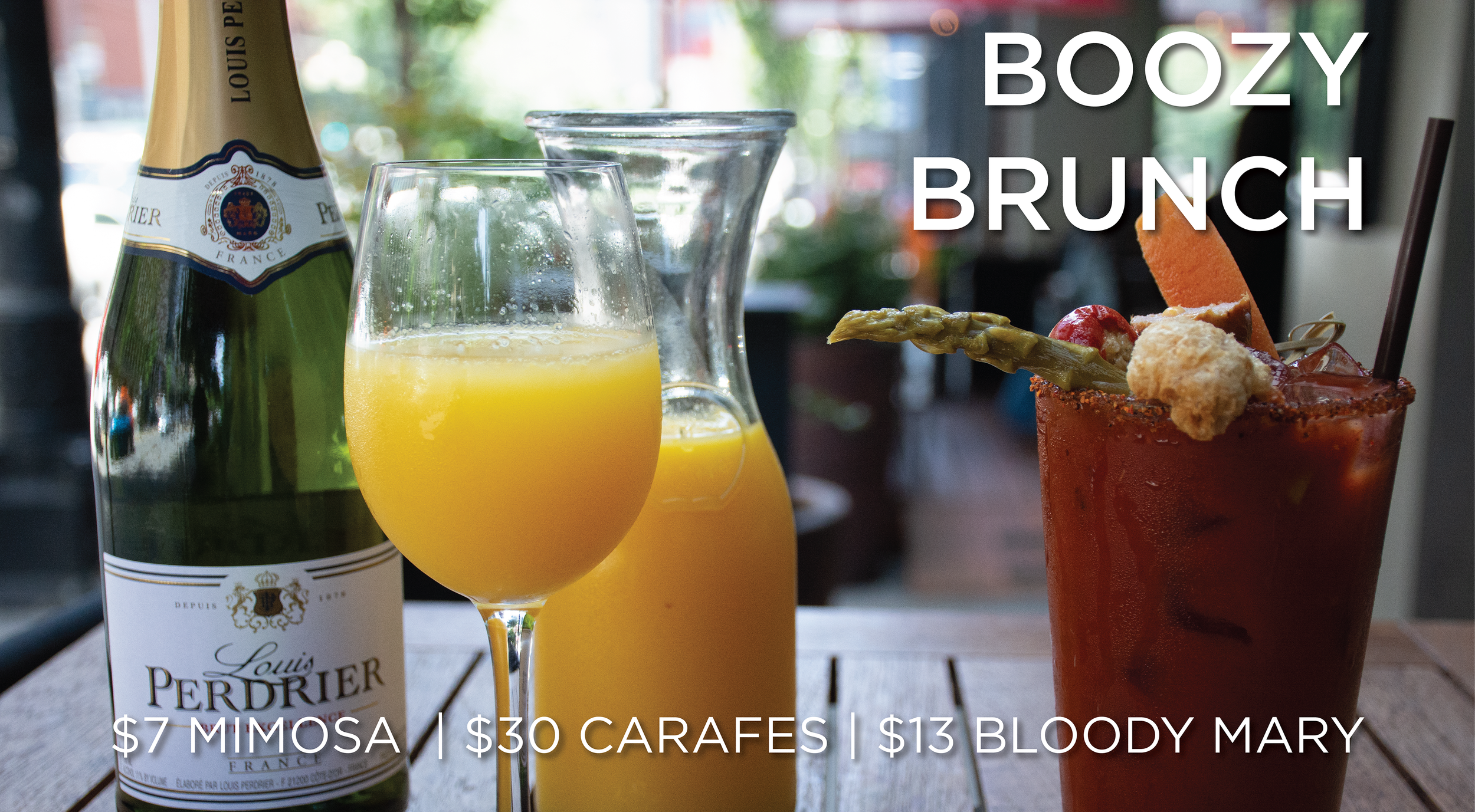 A brunch scene with a bottle of Louis Perdrier Champagne, a glass of orange mimosa, a pitcher of orange juice, and a Bloody Mary garnished with vegetables and a fried snack, set on a wooden table in a restaurant with a blurred outdoor view.