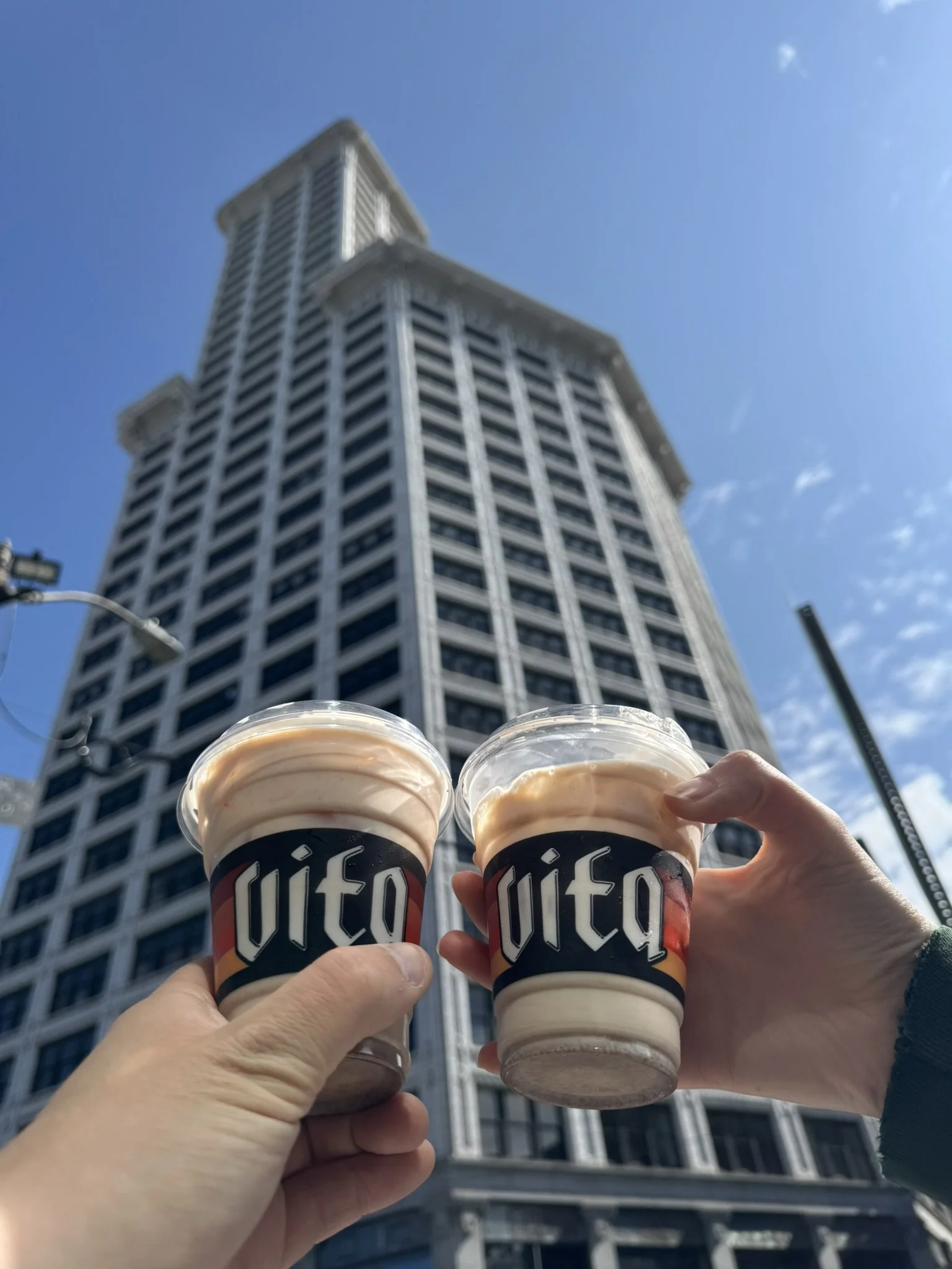 Two hands holding cups of coffee in front of a tall skyscraper on a clear sunny day.