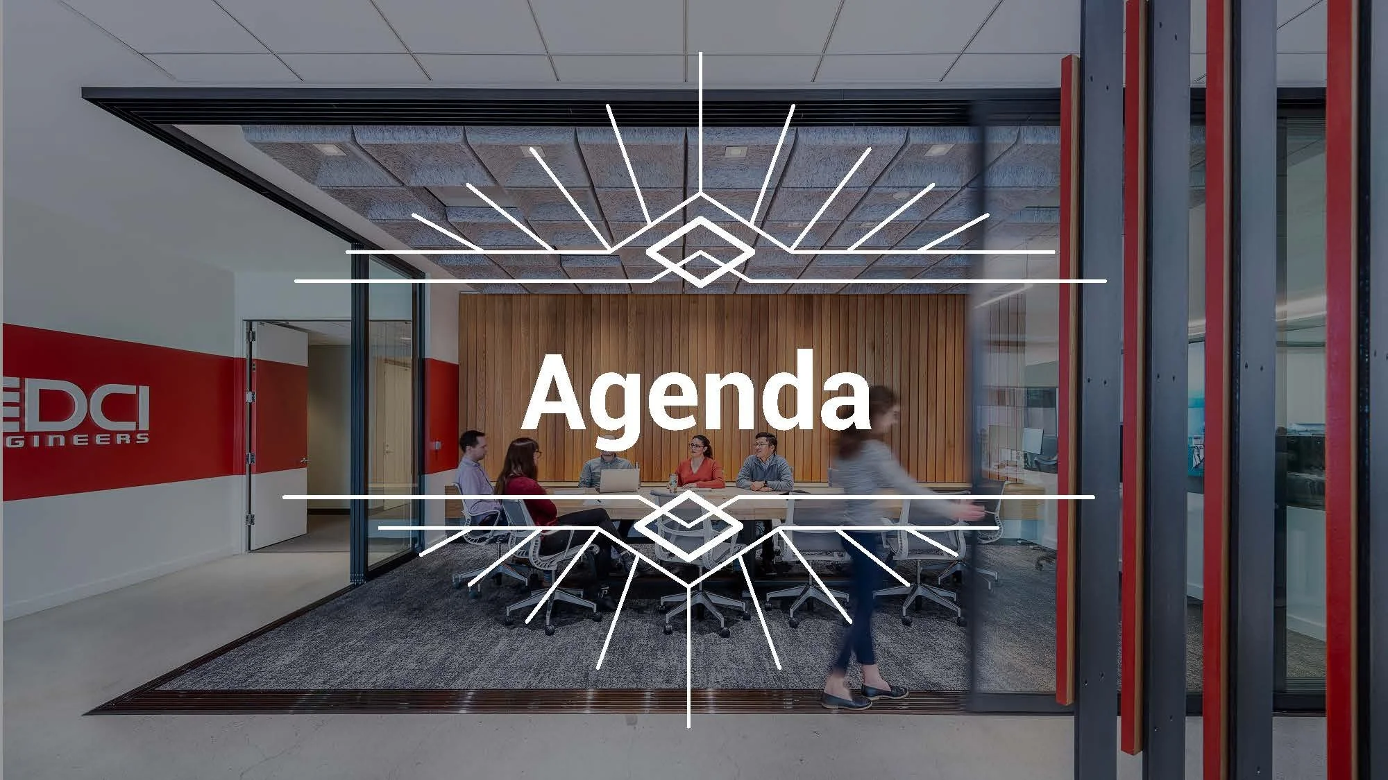 A meeting room with six people sitting around a conference table and one person walking past. The room has wood paneling on the back wall and a modern ceiling light fixture. The glass wall reveals an office space outside. The word 'Agenda' is overlaid on the image.
