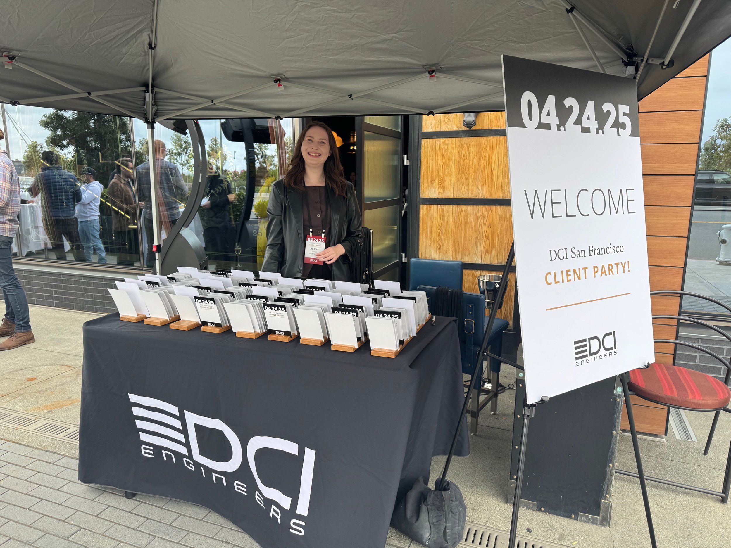 A woman standing behind a table with BRN Engineiers branding, smiling at the camera at an outdoor event. The table has many name badges, with a large welcome sign on a stand next to the table. The event appears to be a client party at DCI San Francisco on April 24, 2025.